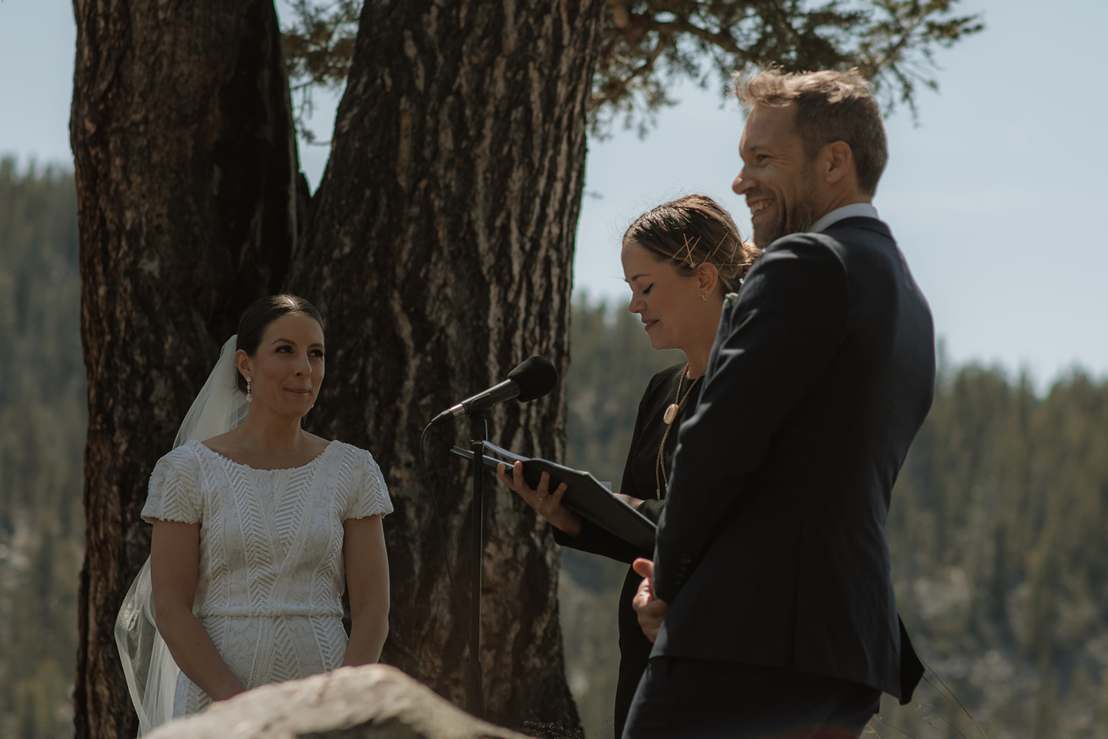 A wedding ceremony taking place outdoors, with a bride and groom standing in front of a large tree as an officiant reads from a book at the wedding tree in Jackson Hole