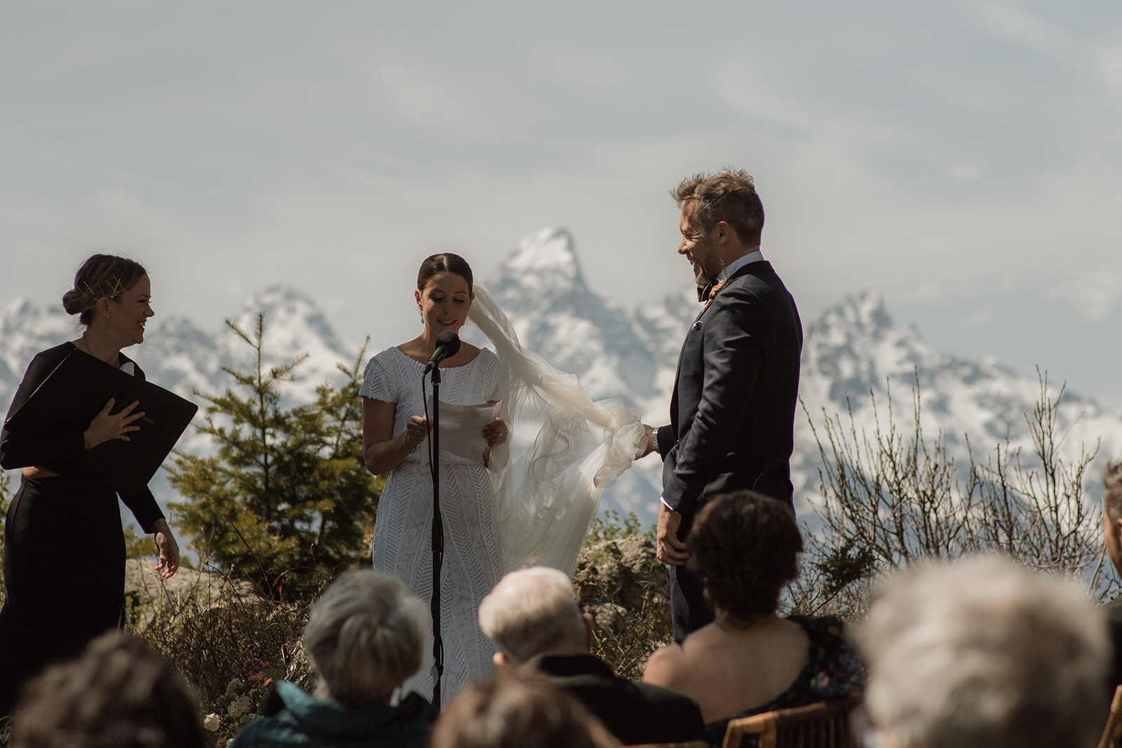 A wedding ceremony taking place outdoors, with a bride and groom standing in front of a large tree as an officiant reads from a book at the wedding tree in Jackson Hole 