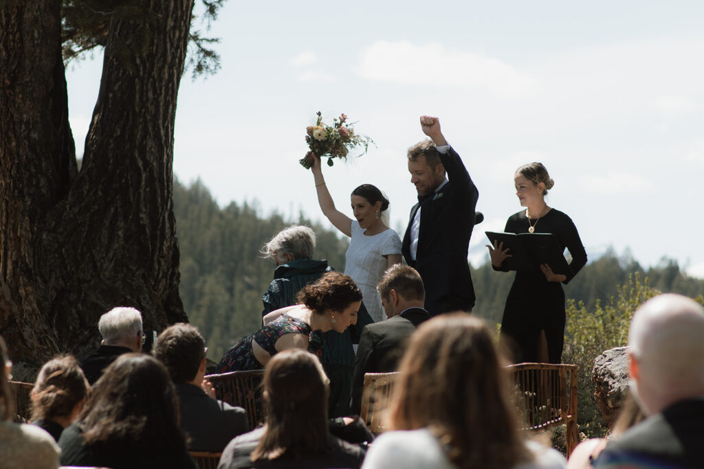 A wedding ceremony taking place outdoors, with a bride and groom standing in front of a large tree as an officiant reads from a book at the wedding tree in Jackson Hole