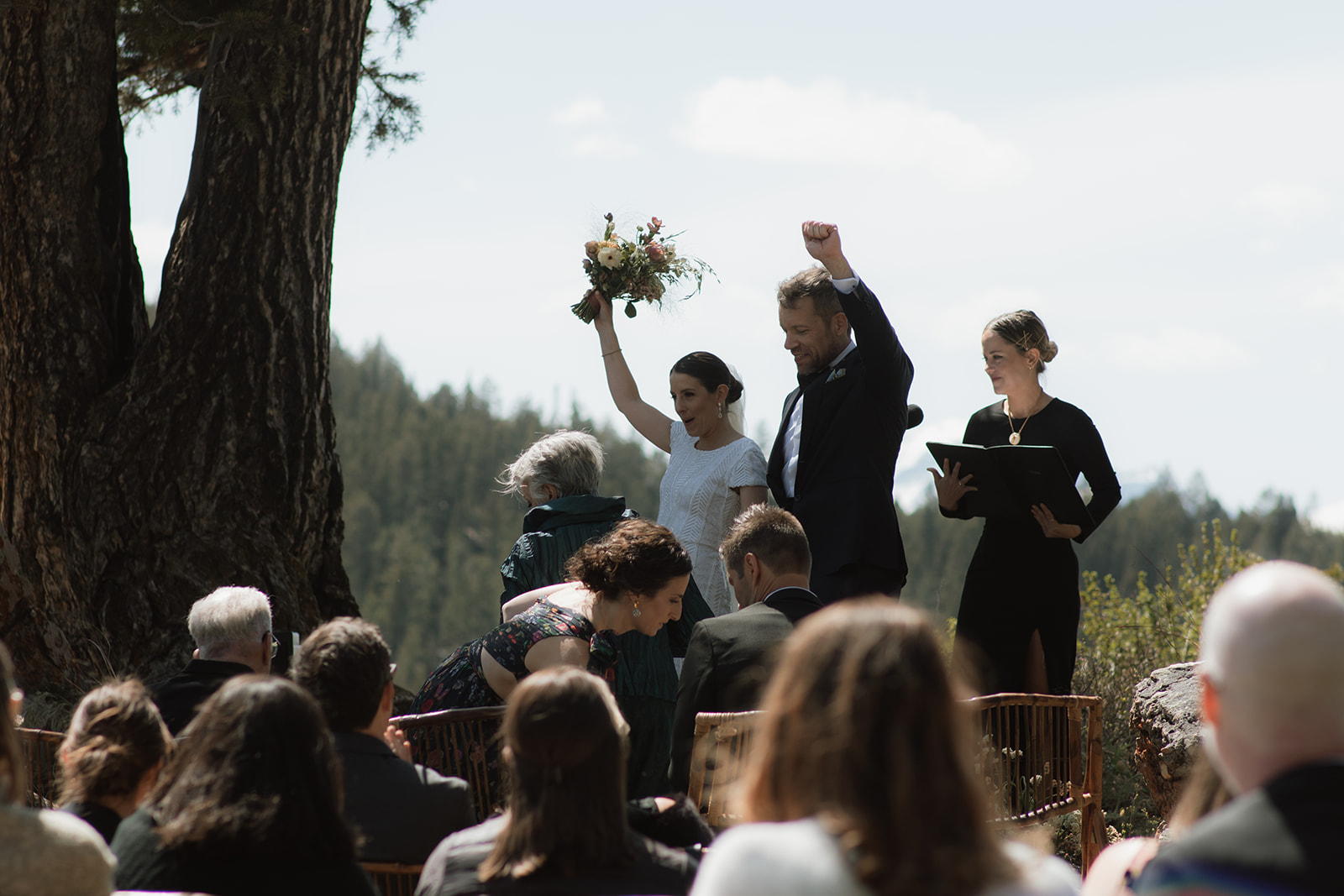 A wedding ceremony taking place outdoors, with a bride and groom standing in front of a large tree as an officiant reads from a book at the wedding tree in Jackson Hole 