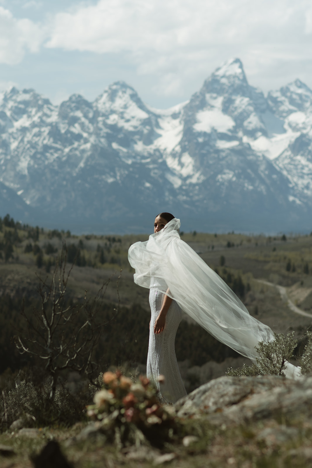 A bride in a white wedding dress and veil stands on a rocky terrain facing snow-capped mountains, with a bouquet of flowers in the foreground at the wedding tree in Jackson Hole