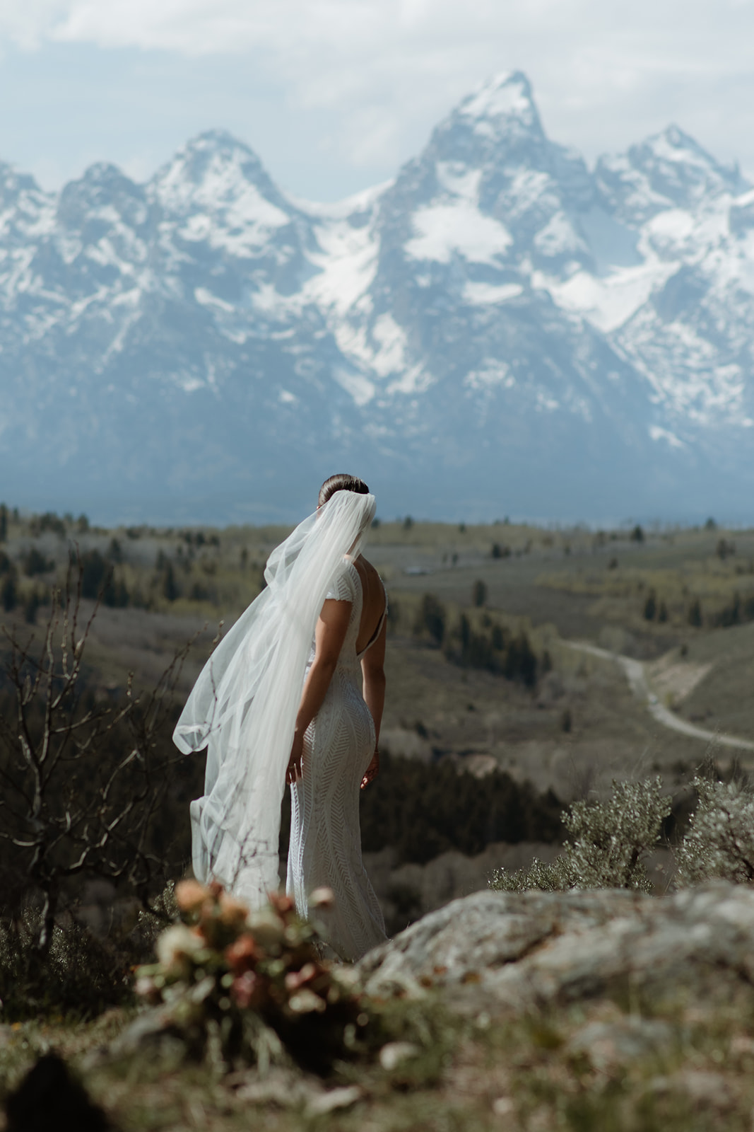 A bride in a white wedding dress and veil stands on a rocky terrain facing snow-capped mountains, with a bouquet of flowers in the foreground at the wedding tree in Jackson Hole