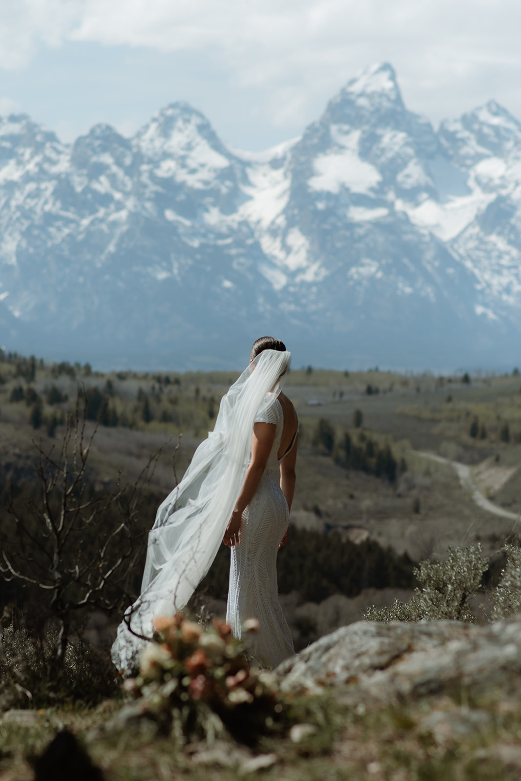 A bride in a white wedding dress and veil stands on a rocky terrain facing snow-capped mountains, with a bouquet of flowers in the foreground at the wedding tree in Jackson Hole