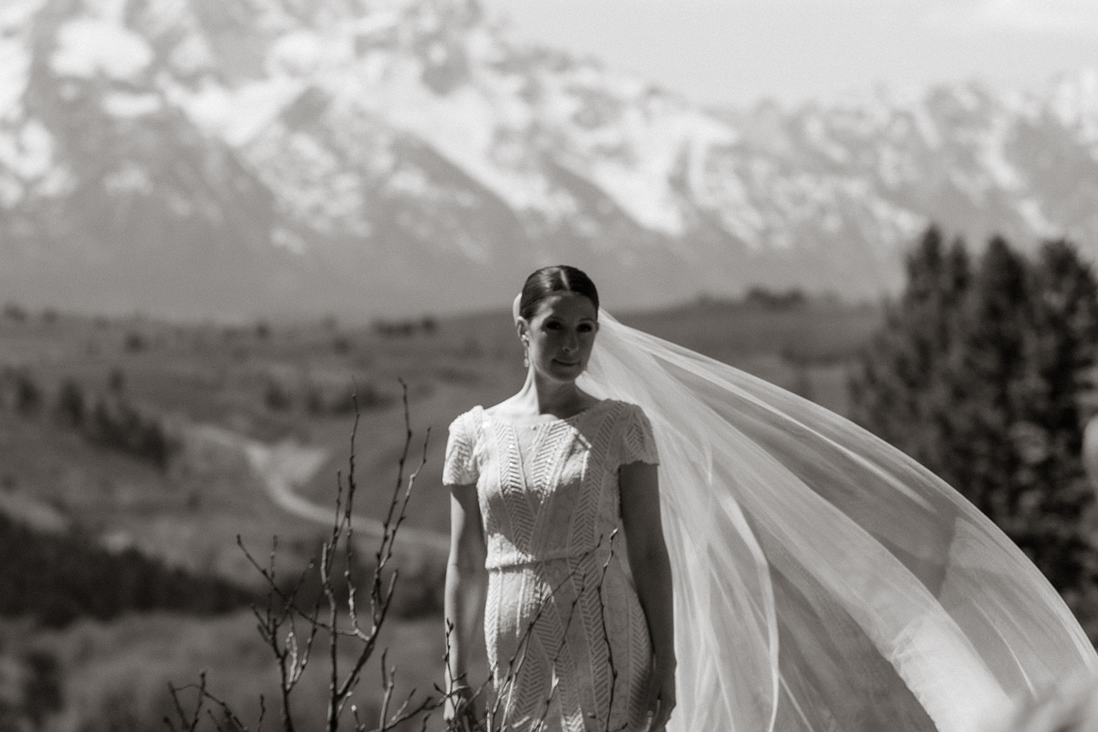 A bride in a white wedding dress and veil stands on a rocky terrain facing snow-capped mountains, with a bouquet of flowers in the foreground at the wedding tree in Jackson Hole