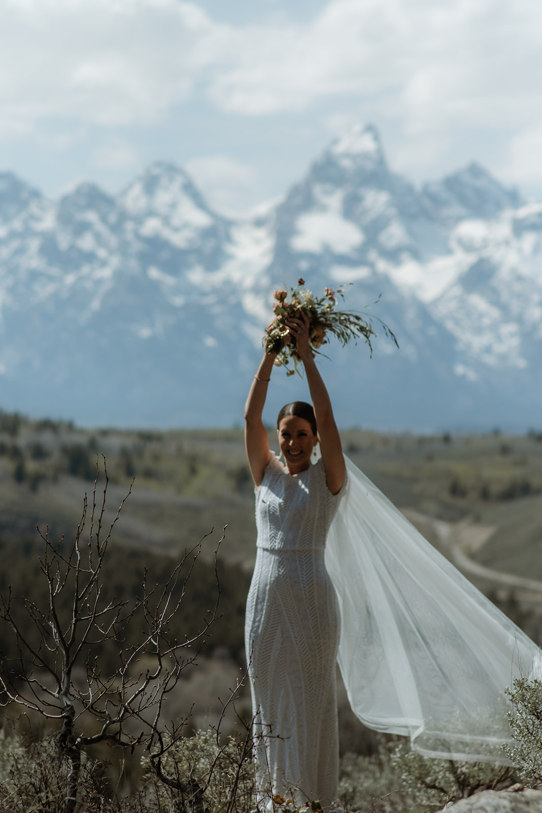 A bride in a white wedding dress and veil stands on a rocky terrain facing snow-capped mountains, with a bouquet of flowers in the foreground at the wedding tree in Jackson Hole