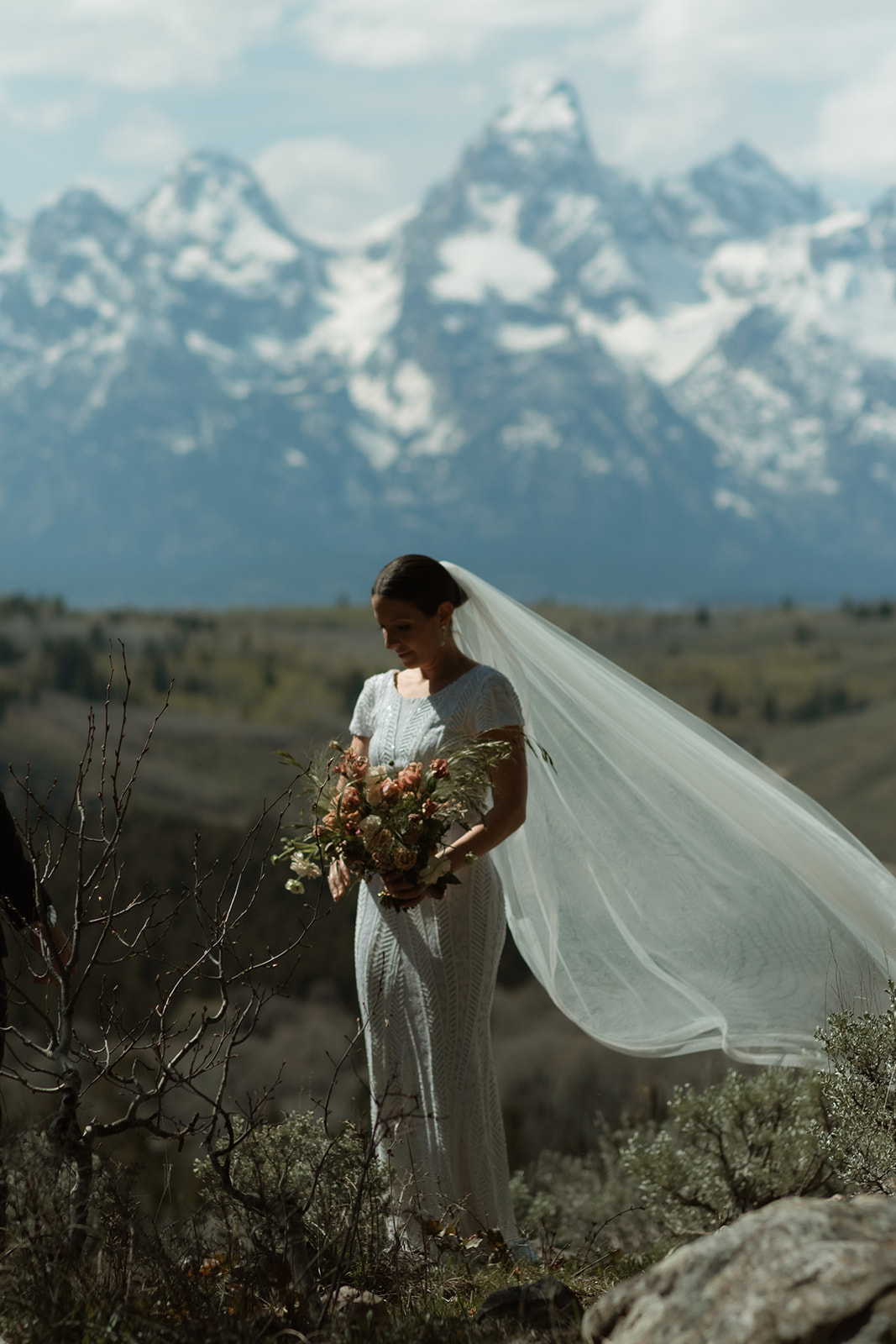 A bride in a white wedding dress and veil stands on a rocky terrain facing snow-capped mountains, with a bouquet of flowers in the foreground at the wedding tree in Jackson Hole