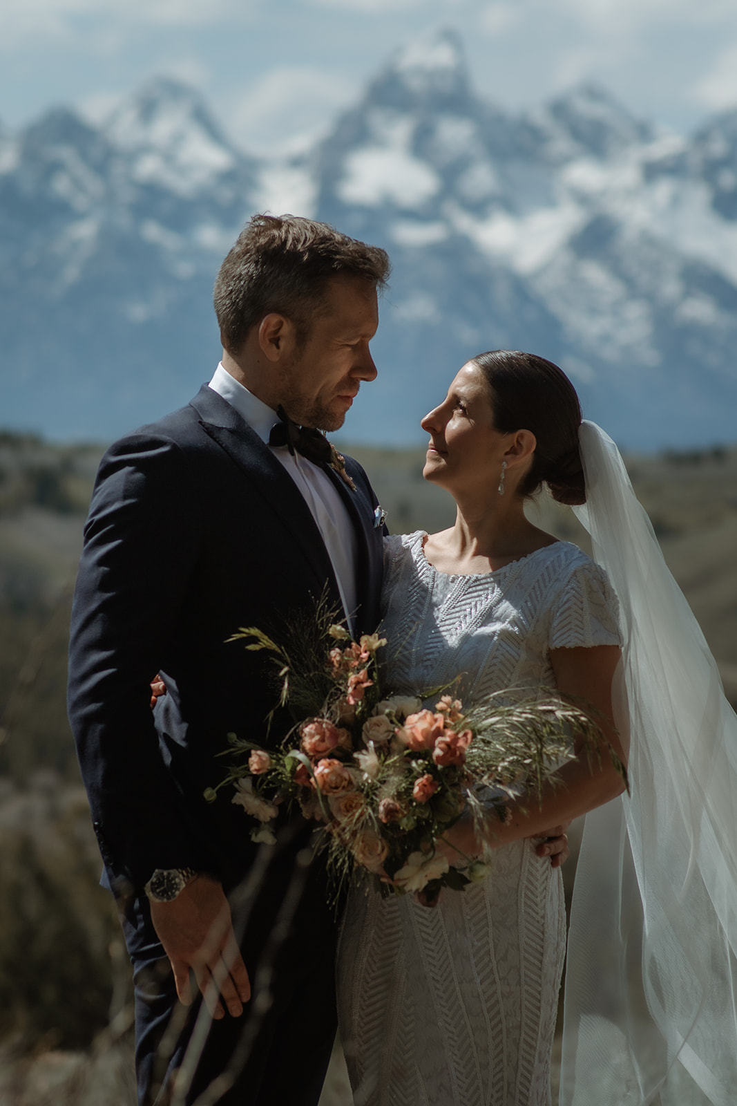 A bride and groom gaze at each other while standing outdoors with snow-capped mountains in the background. The bride holds a bouquet of flowers at the wedding tree in jackson hole