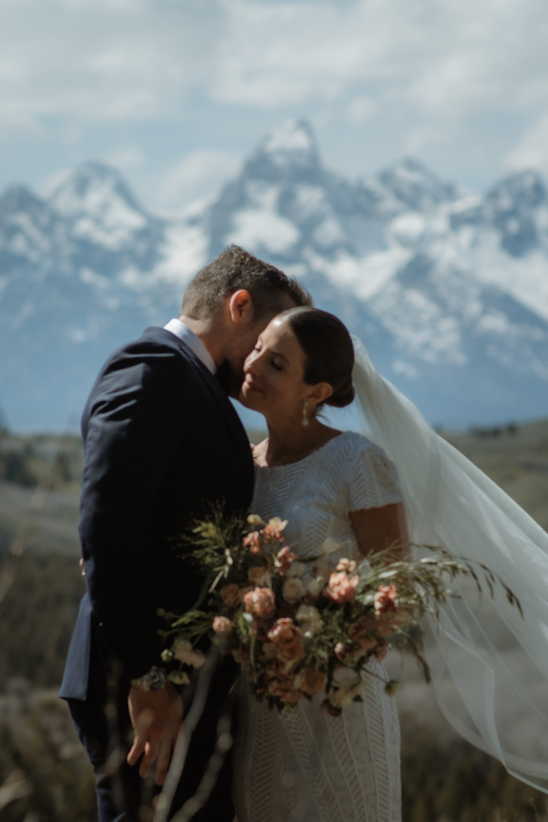 A bride and groom gaze at each other while standing outdoors with snow-capped mountains in the background. The bride holds a bouquet of flowers at the wedding tree in jackson hole