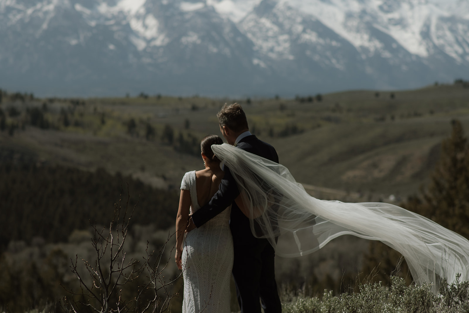 A bride and groom gaze at each other while standing outdoors with snow-capped mountains in the background. The bride holds a bouquet of flowers at the wedding tree in jackson hole