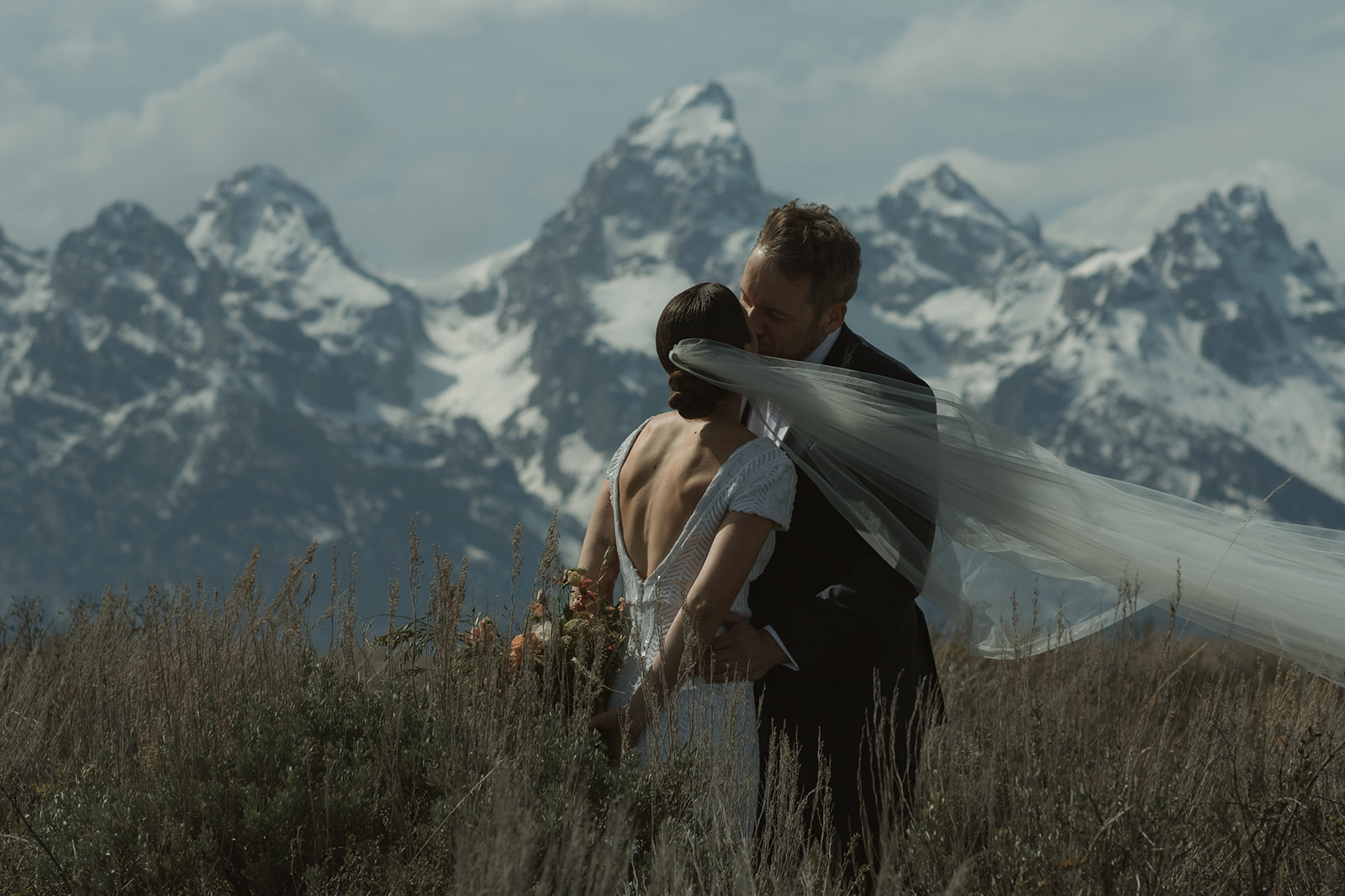 A bride and groom gaze at each other while standing outdoors with snow-capped mountains in the background. The bride holds a bouquet of flowers at the wedding tree in jackson hole