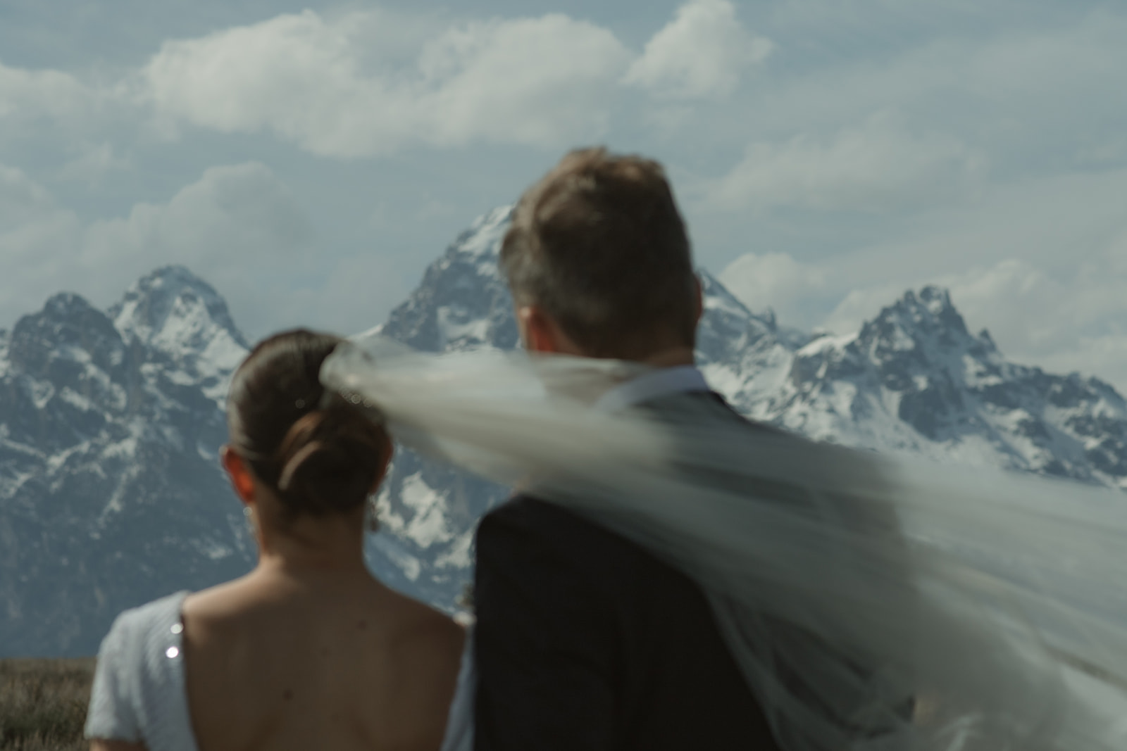 A bride and groom gaze at each other while standing outdoors with snow-capped mountains in the background. The bride holds a bouquet of flowers 