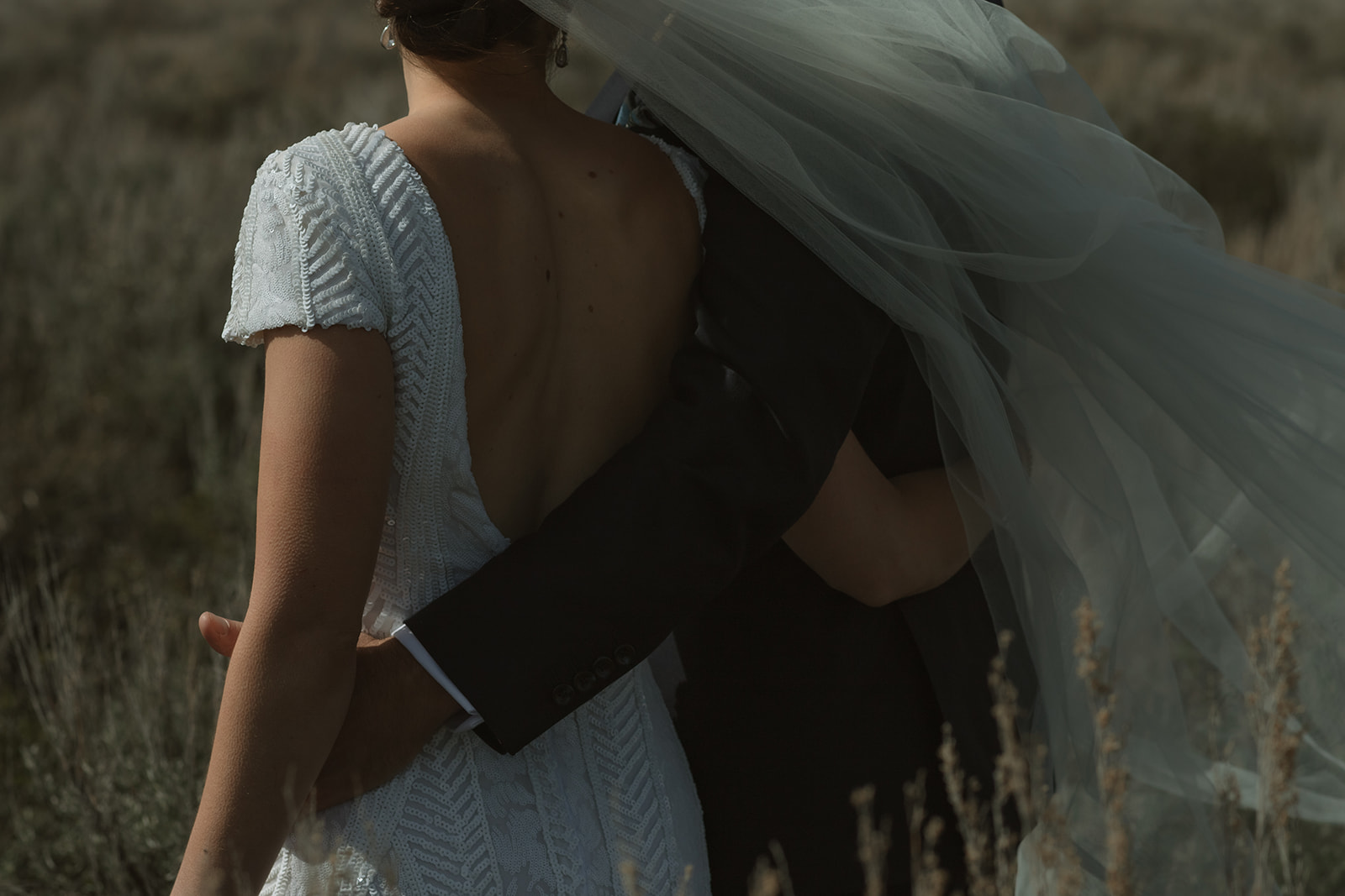 A bride and groom gaze at each other while standing outdoors with snow-capped mountains in the background. The bride holds a bouquet of flowers 