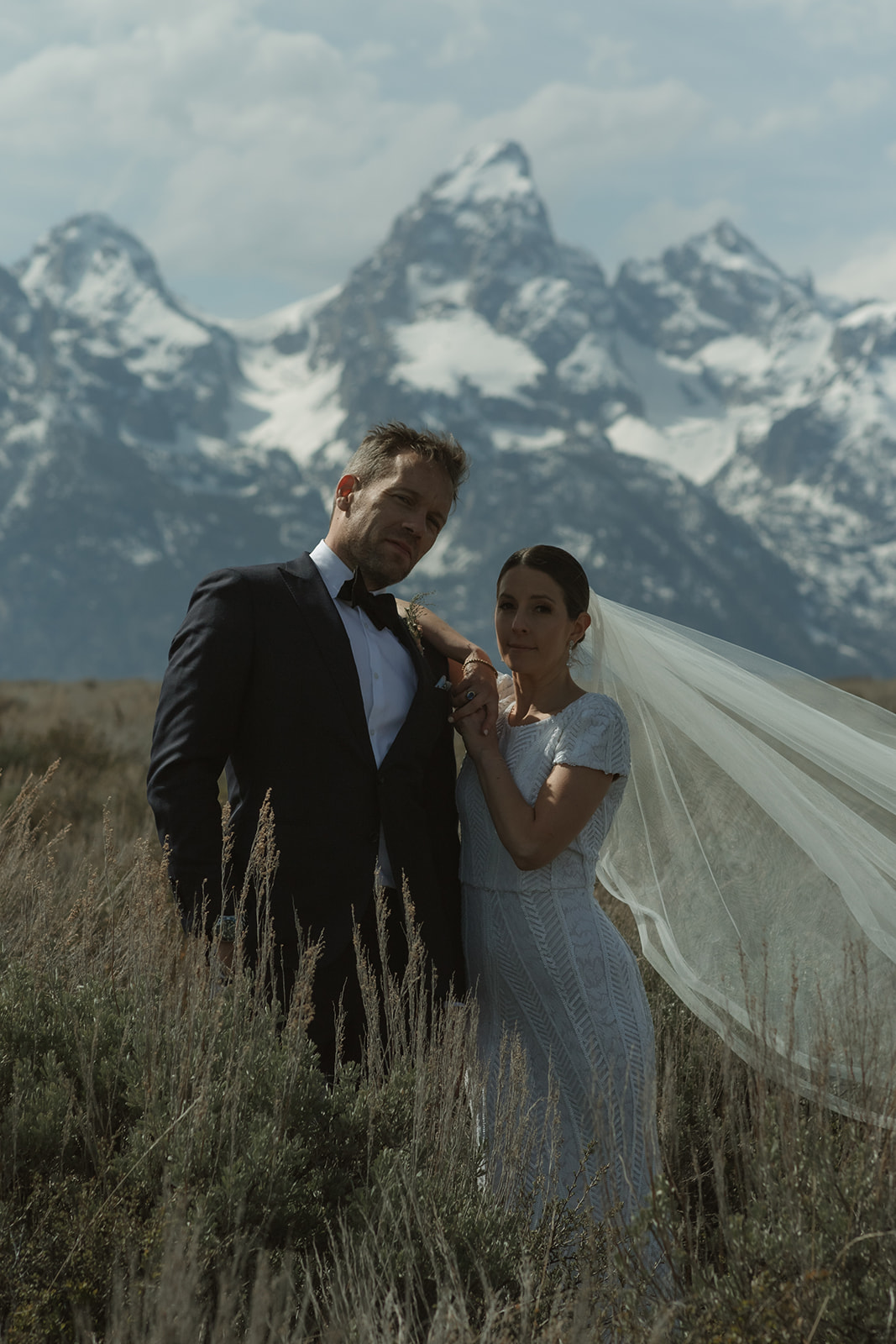 A bride and groom gaze at each other while standing outdoors with snow-capped mountains in the background. The bride holds a bouquet of flowers 