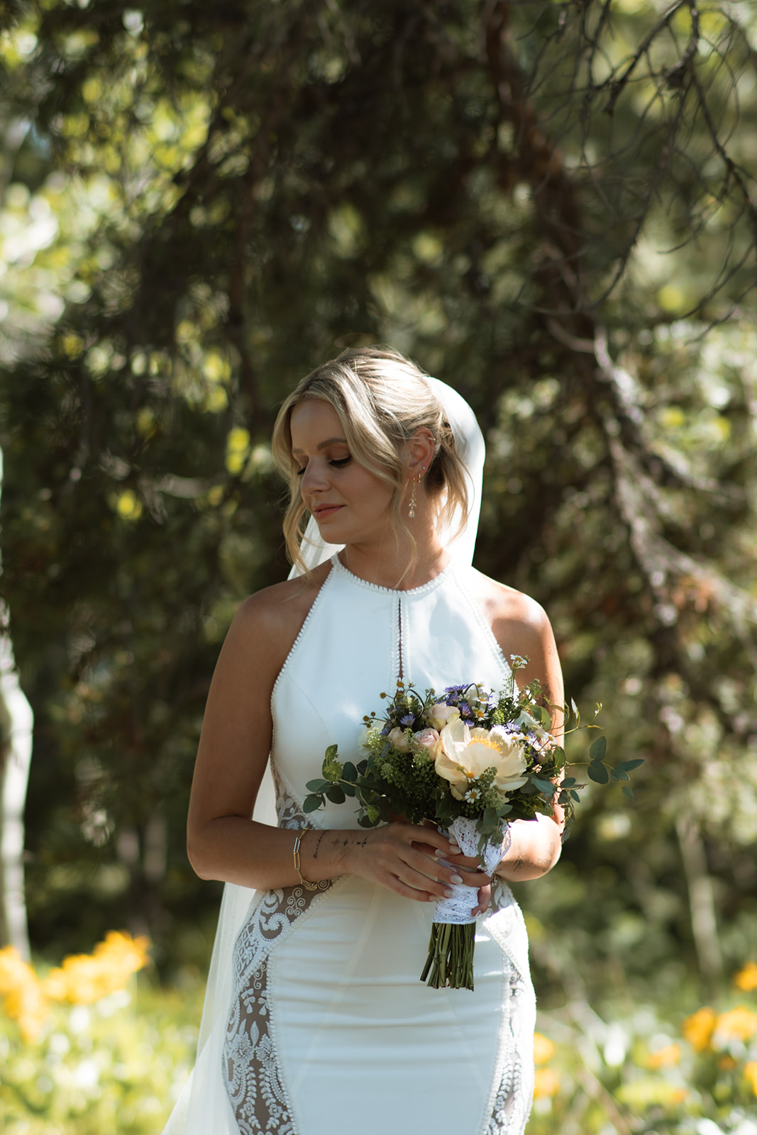 A bride in a white sleeveless wedding dress holds a bouquet of flowers while standing outdoors in a forested area during an elopement at Colter bay at Jackson Hole