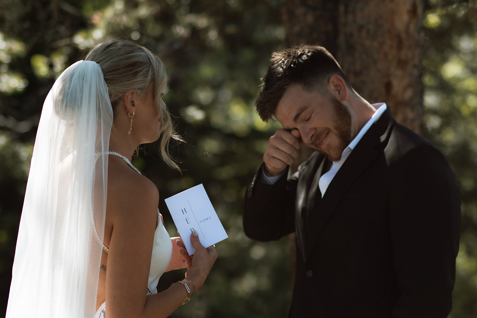 A bride and groom stand outdoors by a tree. The groom, in a black suit, reads from a piece of paper while the bride, in a white dress with a long veil, stands facing him at their JACKSON HOLE ELOPEMENT at Colter Bay