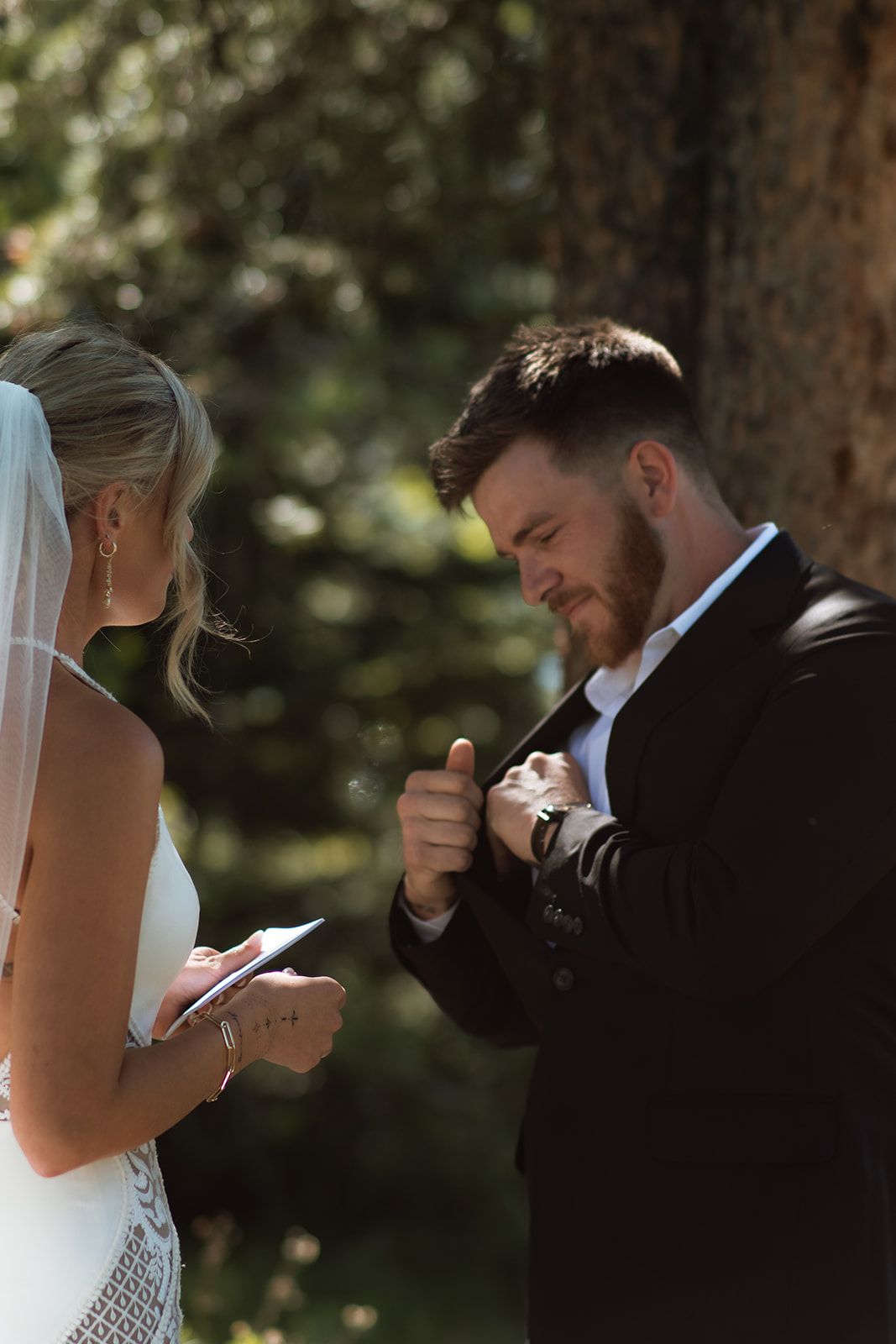 A bride and groom stand outdoors by a tree. The groom, in a black suit, reads from a piece of paper while the bride, in a white dress with a long veil, stands facing him at their JACKSON HOLE ELOPEMENT at Colter Bay