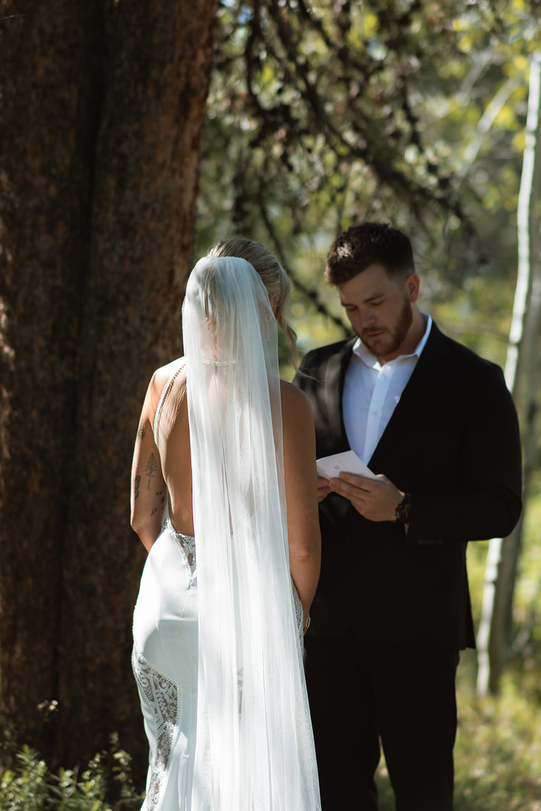 A bride and groom stand outdoors by a tree. The groom, in a black suit, reads from a piece of paper while the bride, in a white dress with a long veil, stands facing him at their JACKSON HOLE ELOPEMENT at Colter Bay