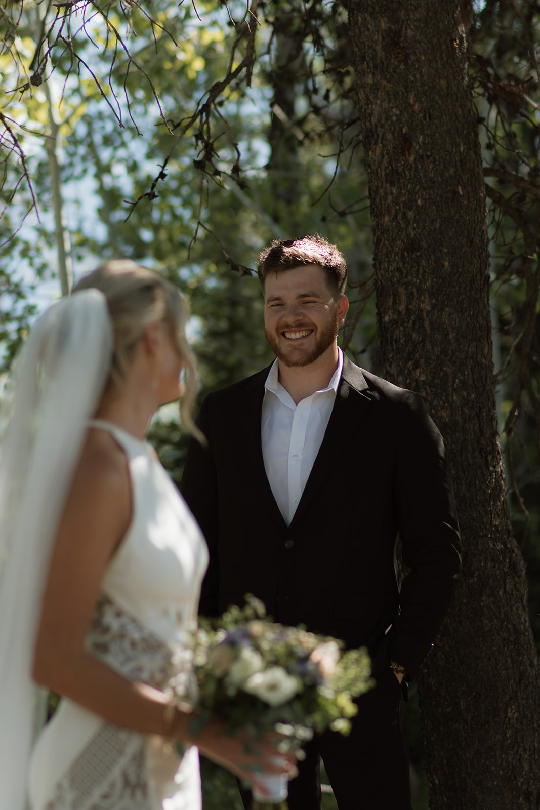 A bride and groom stand outdoors by a tree. The groom, in a black suit, reads from a piece of paper while the bride, in a white dress with a long veil, stands facing him at their JACKSON HOLE ELOPEMENT at Colter Bay