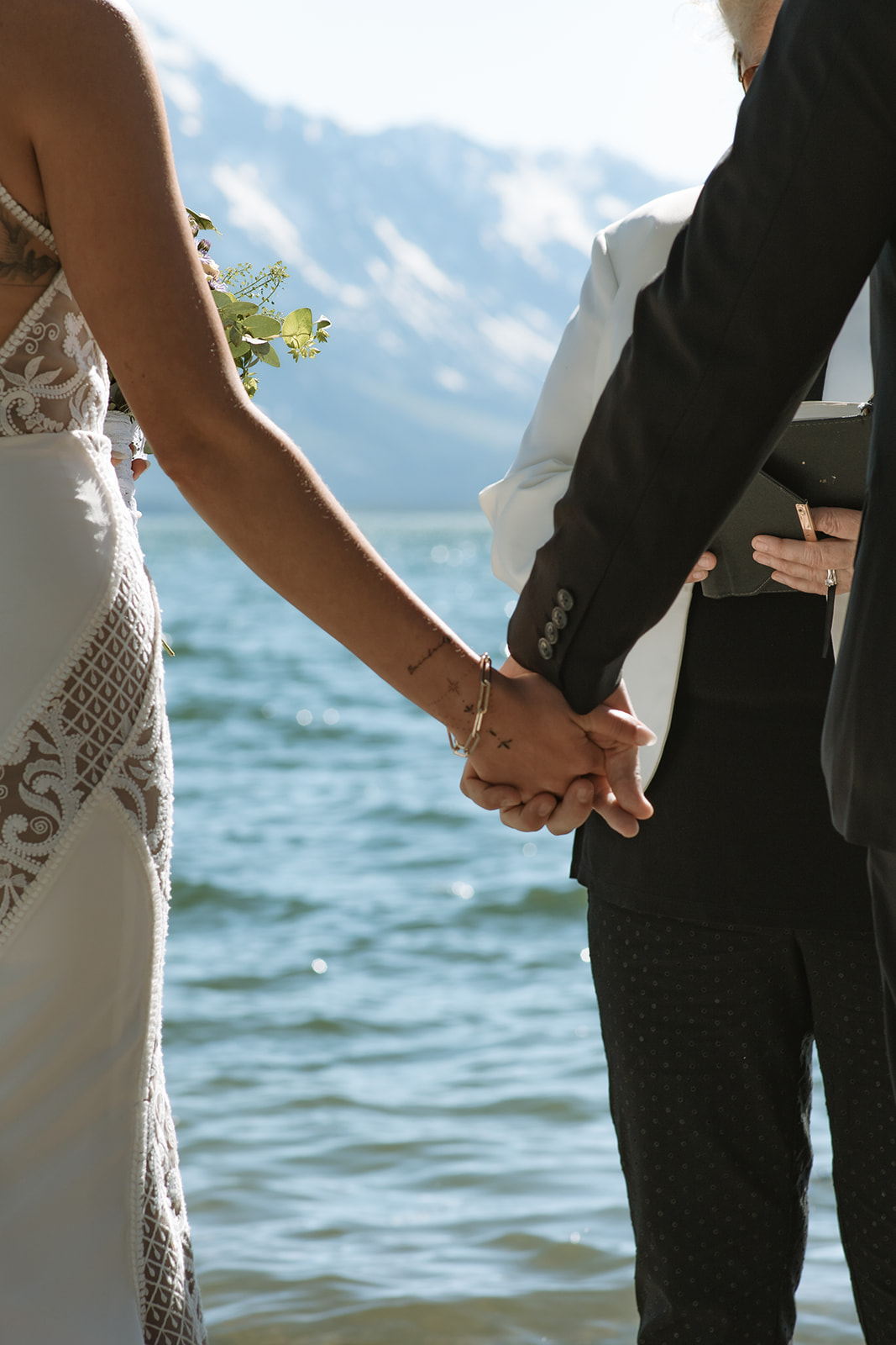 A couple exchanges rings in a wedding ceremony by a body of water, officiated by a woman holding a book. The bride is in a white dress with a veil, and the groom is in a dark suit at an elopement at Colter Bay in Jackson Hole 