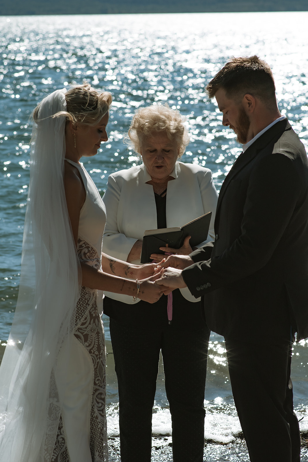 A couple exchanges rings in a wedding ceremony by a body of water, officiated by a woman holding a book. The bride is in a white dress with a veil, and the groom is in a dark suit at an elopement at Colter Bay in Jackson Hole 