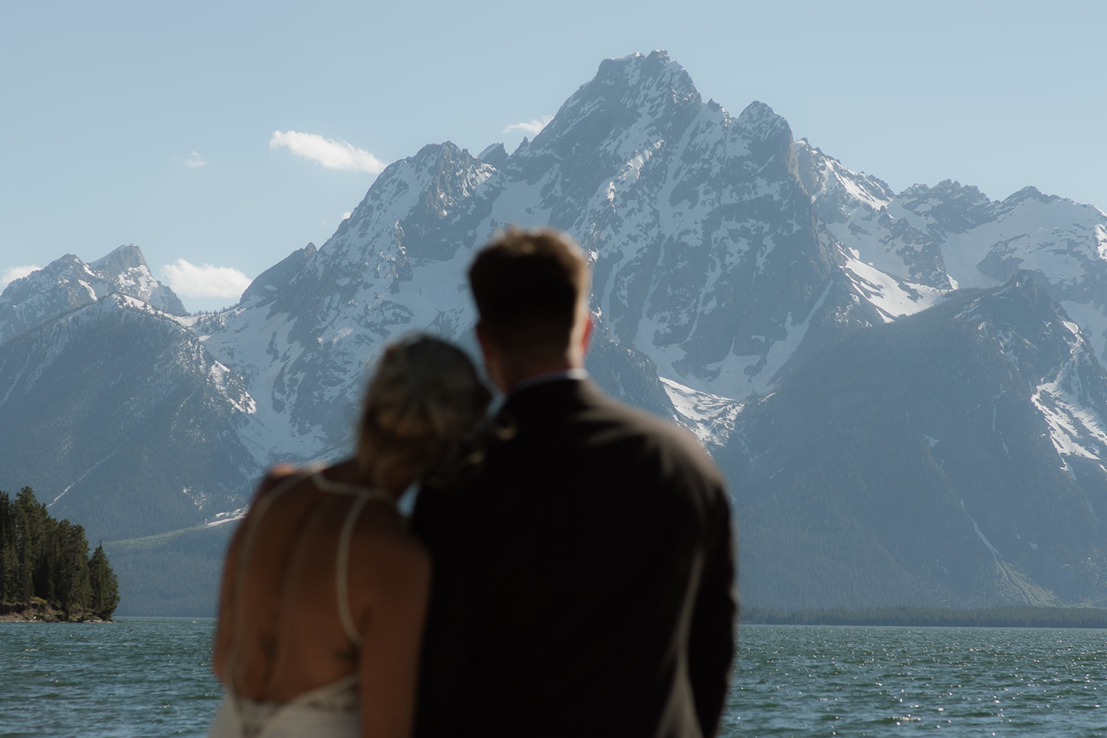 A couple stands close, nearly touching noses, with a snowy mountain range in the background. The woman wears a sleeveless dress, and the man is in a dark suit for their jackson hole elopement