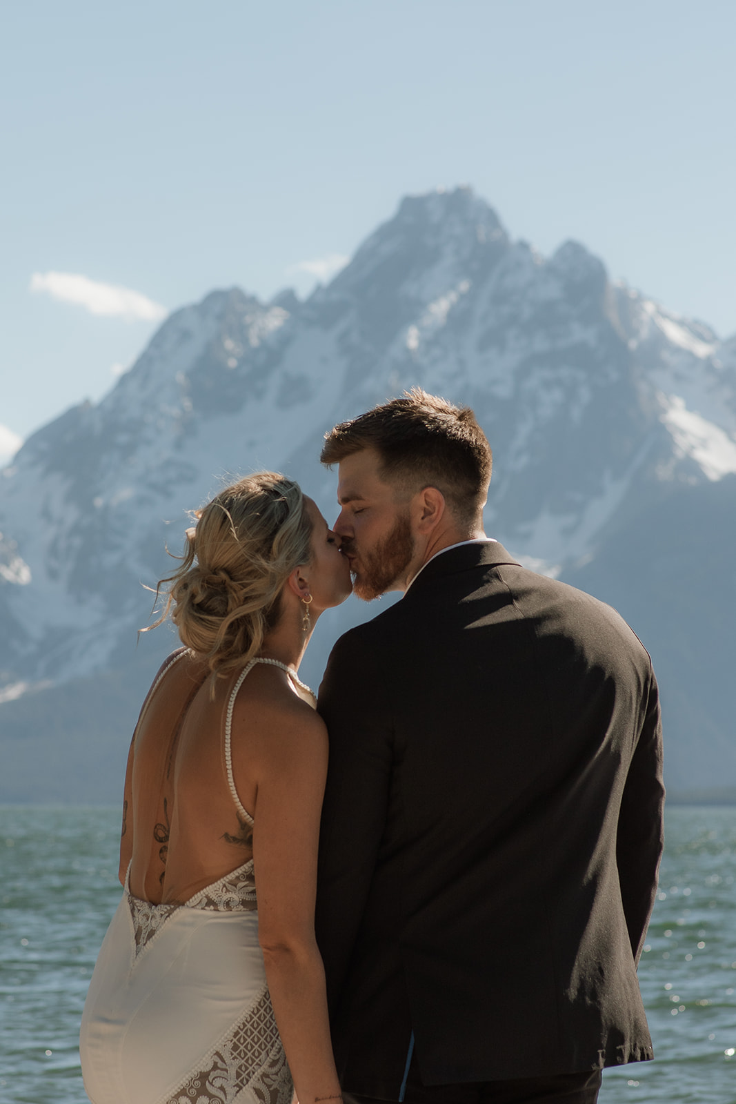 A couple stands close, nearly touching noses, with a snowy mountain range in the background. The woman wears a sleeveless dress, and the man is in a dark suit for their jackson hole elopement 