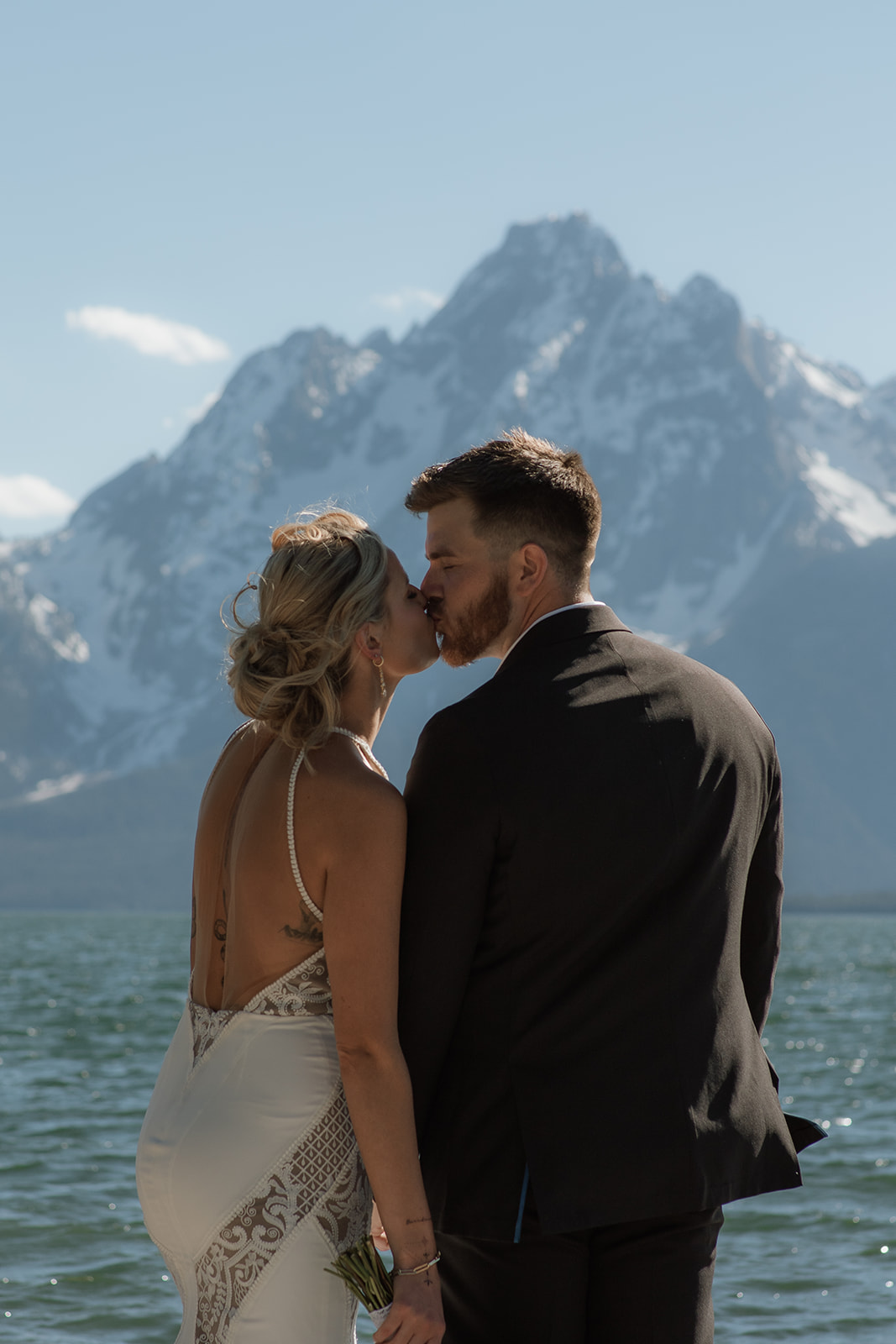 A couple dressed in wedding attire kisses by a lake with snowy mountains in the background.