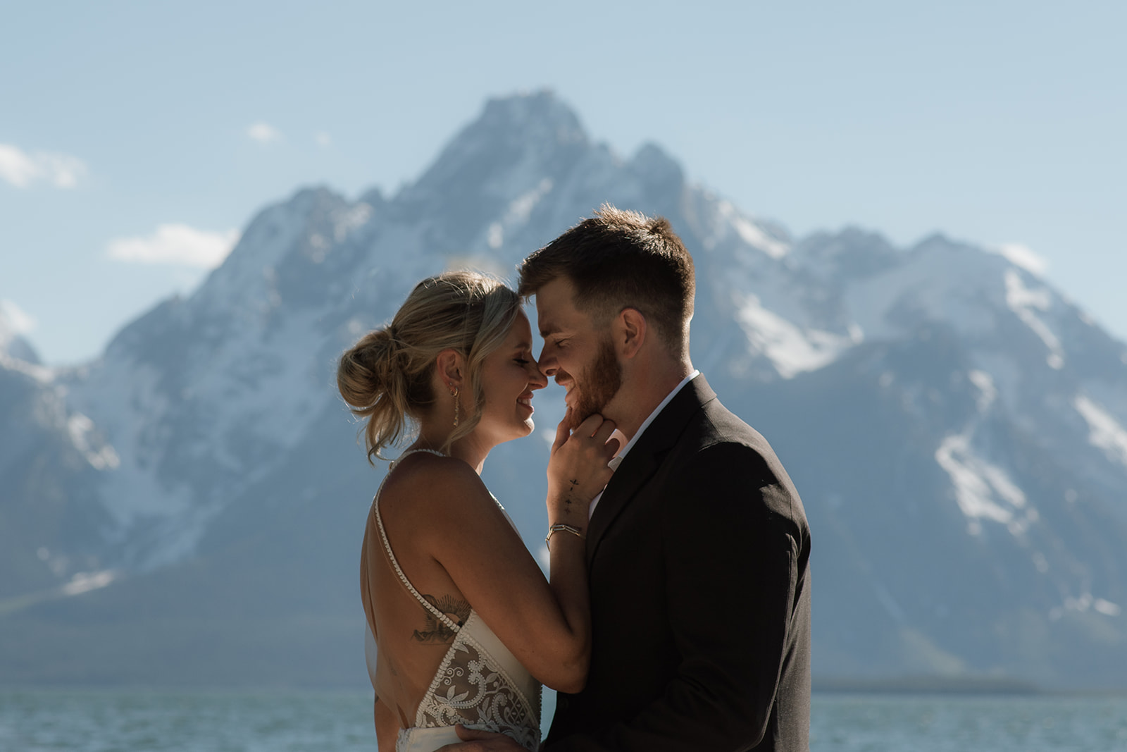 A couple stands close, nearly touching noses, with a snowy mountain range in the background. The woman wears a sleeveless dress, and the man is in a dark suit for their jackson hole elopement