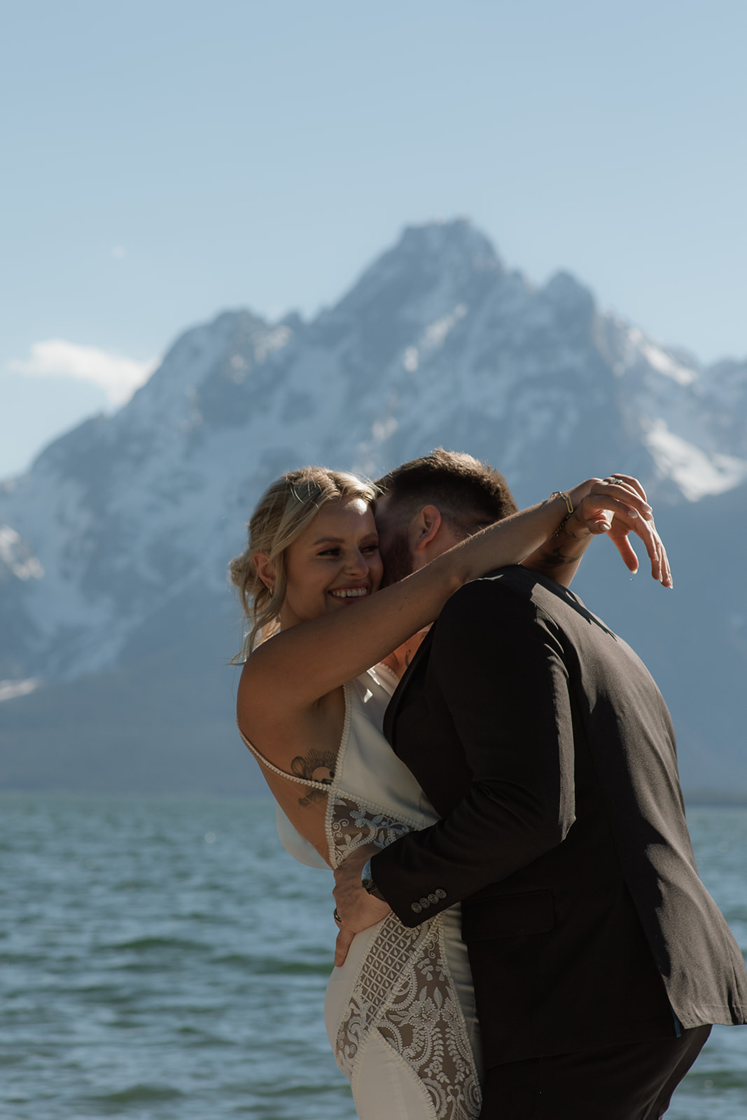 A couple stands close, nearly touching noses, with a snowy mountain range in the background. The woman wears a sleeveless dress, and the man is in a dark suit for their jackson hole elopement