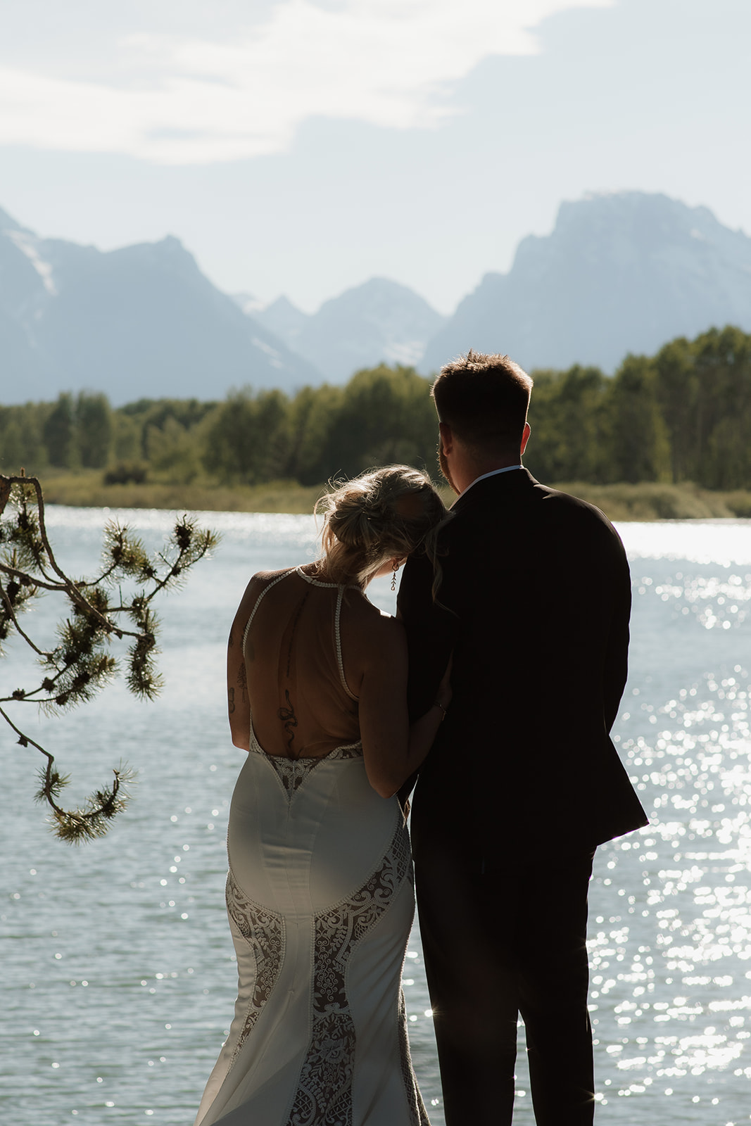 A couple stands by a lake, facing away, with mountains and trees in the background under a partly cloudy sky. She is wearing a white dress, and he is in a black suitfor their elopement at jackson hole 