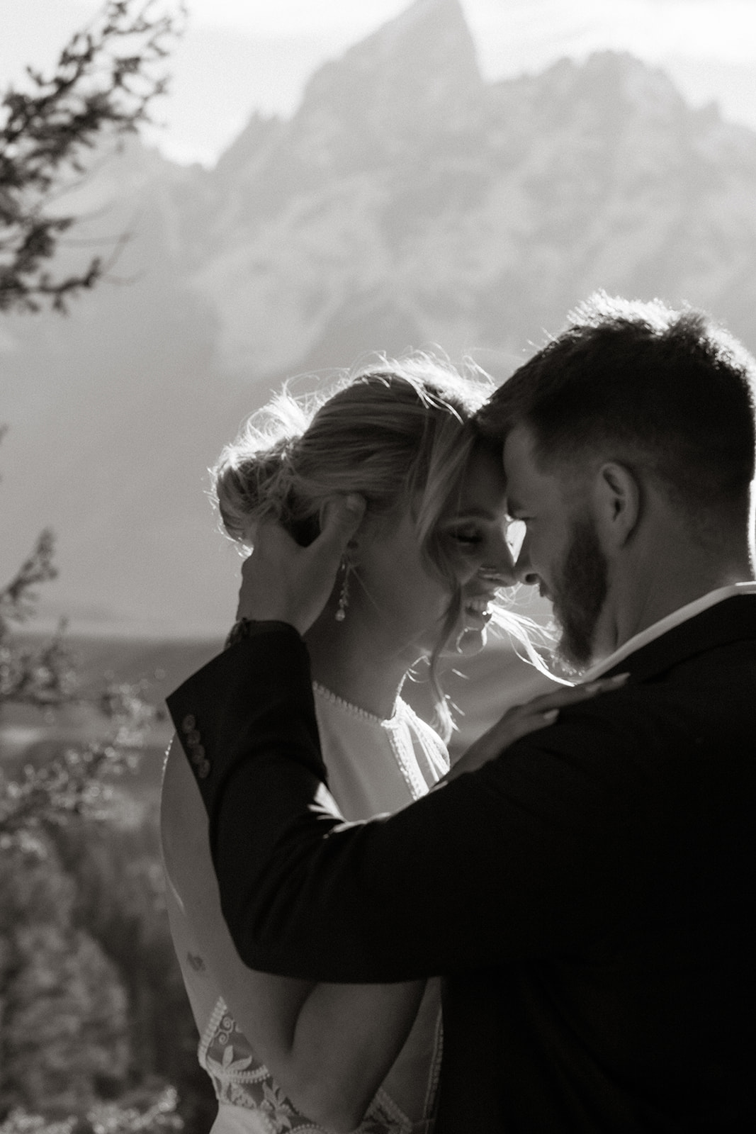 A couple stands by a lake, facing away, with mountains and trees in the background under a partly cloudy sky. She is wearing a white dress, and he is in a black suit for their elopement at jackson hole 