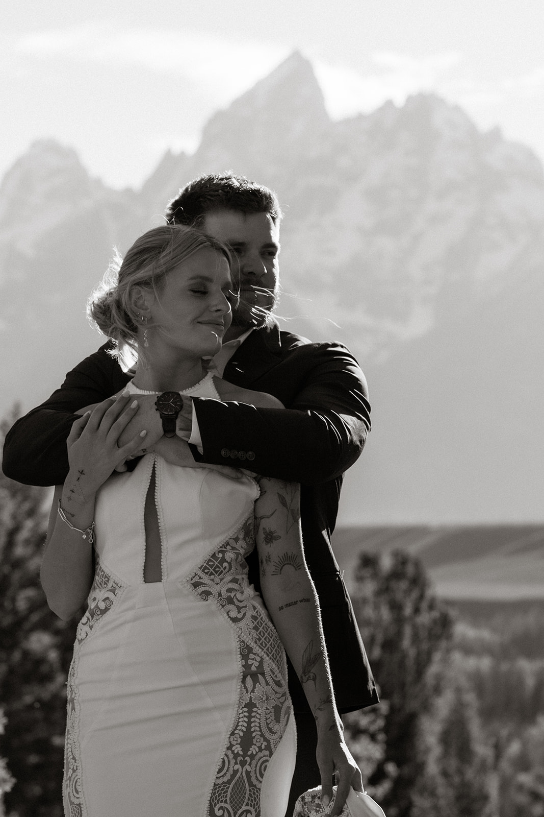 A couple stands by a lake, facing away, with mountains and trees in the background under a partly cloudy sky. She is wearing a white dress, and he is in a black suit for their elopement at jackson hole 