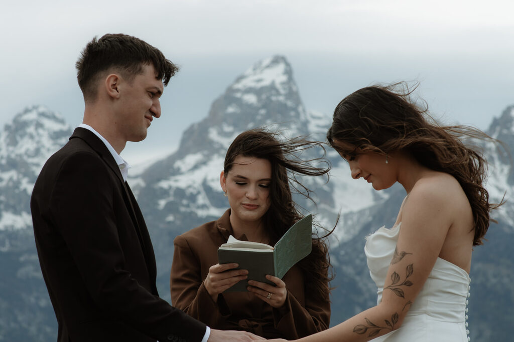 A couple stands facing each other holding hands during a wedding ceremony outdoors, with a person in the middle reading from a book. Snow-capped mountains are visible in the background for their elopment at Glacier View Turnout