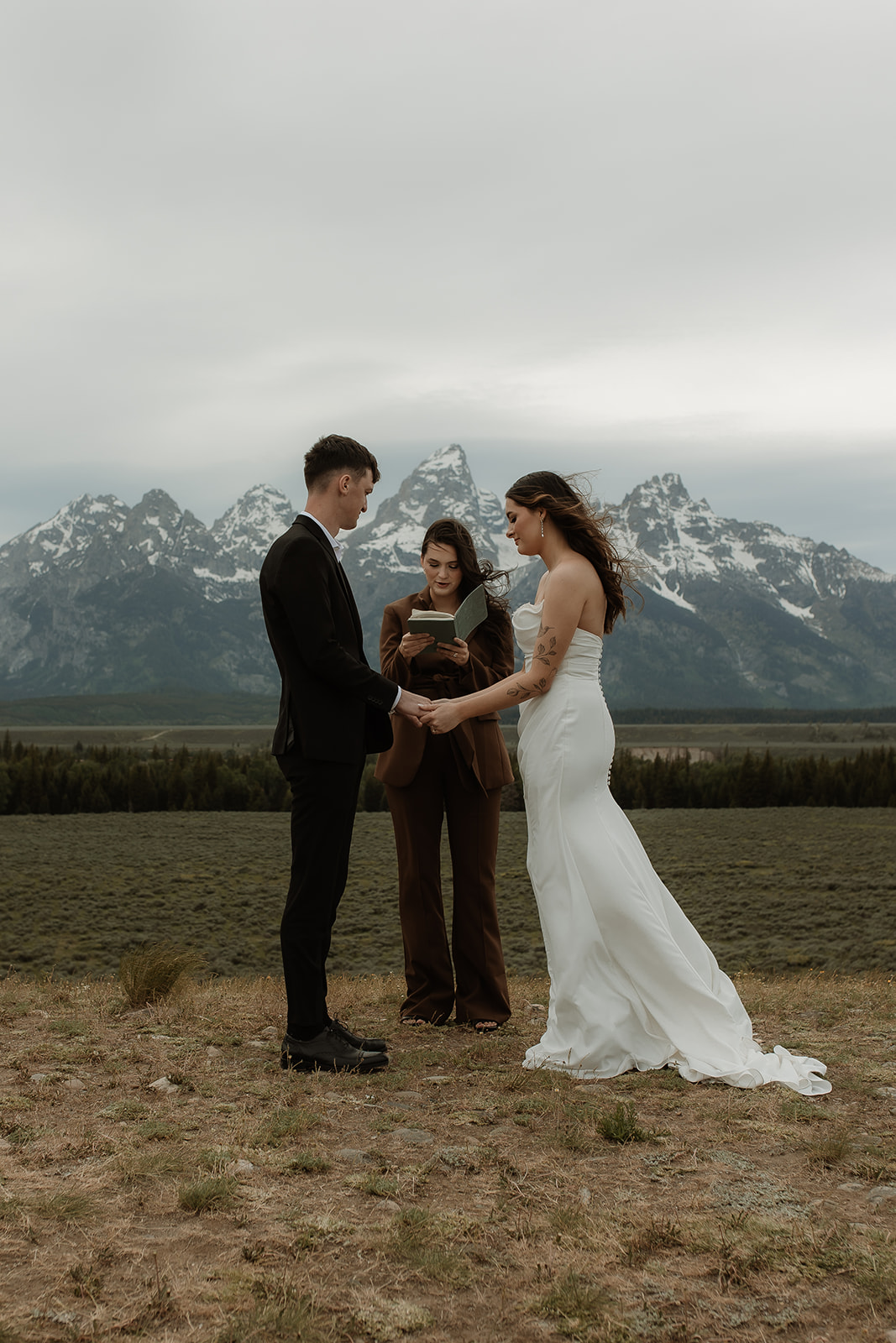 A couple stands facing each other holding hands during a wedding ceremony outdoors, with a person in the middle reading from a book. Snow-capped mountains are visible in the background for their elopment at Glacier View Turnout