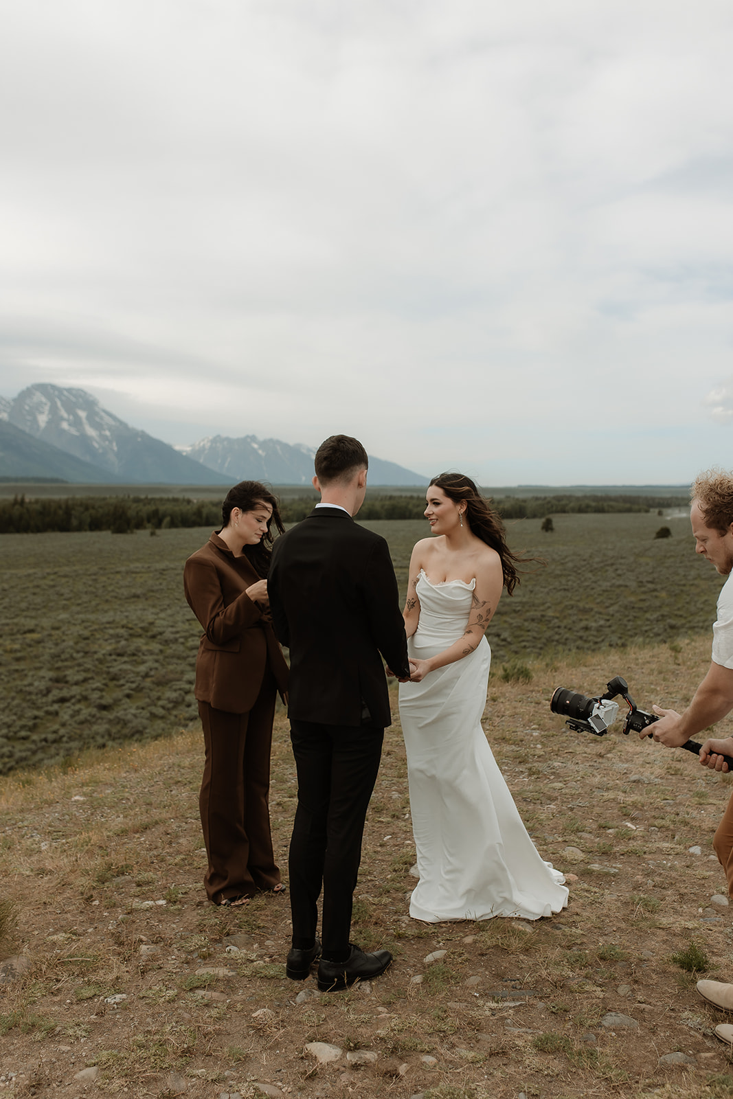 A couple stands facing each other holding hands during a wedding ceremony outdoors, with a person in the middle reading from a book. Snow-capped mountains are visible in the background for their elopment at Glacier View Turnout