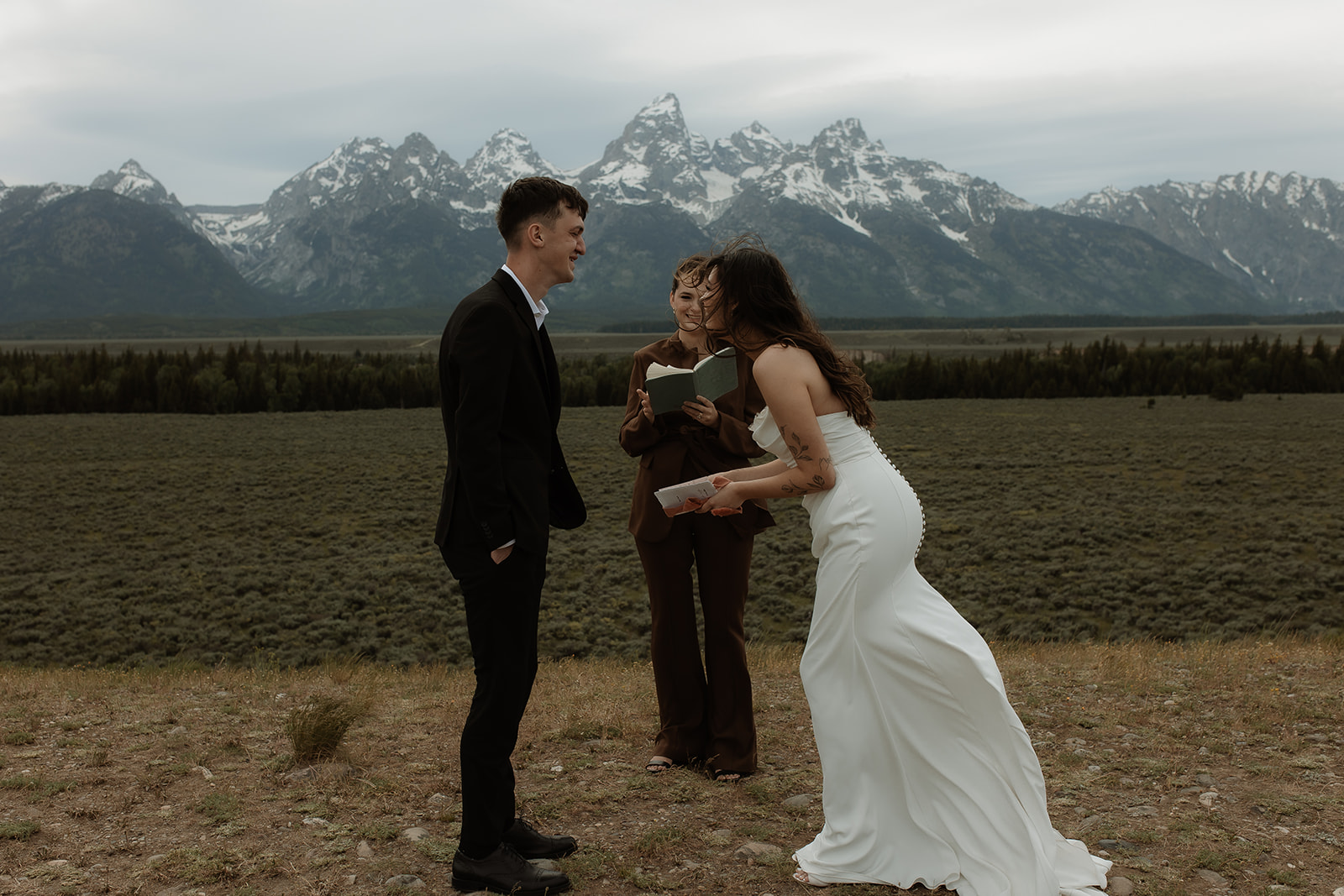 A couple stands facing each other holding hands during a wedding ceremony outdoors, with a person in the middle reading from a book. Snow-capped mountains are visible in the background for their elopment at Glacier View Turnout 