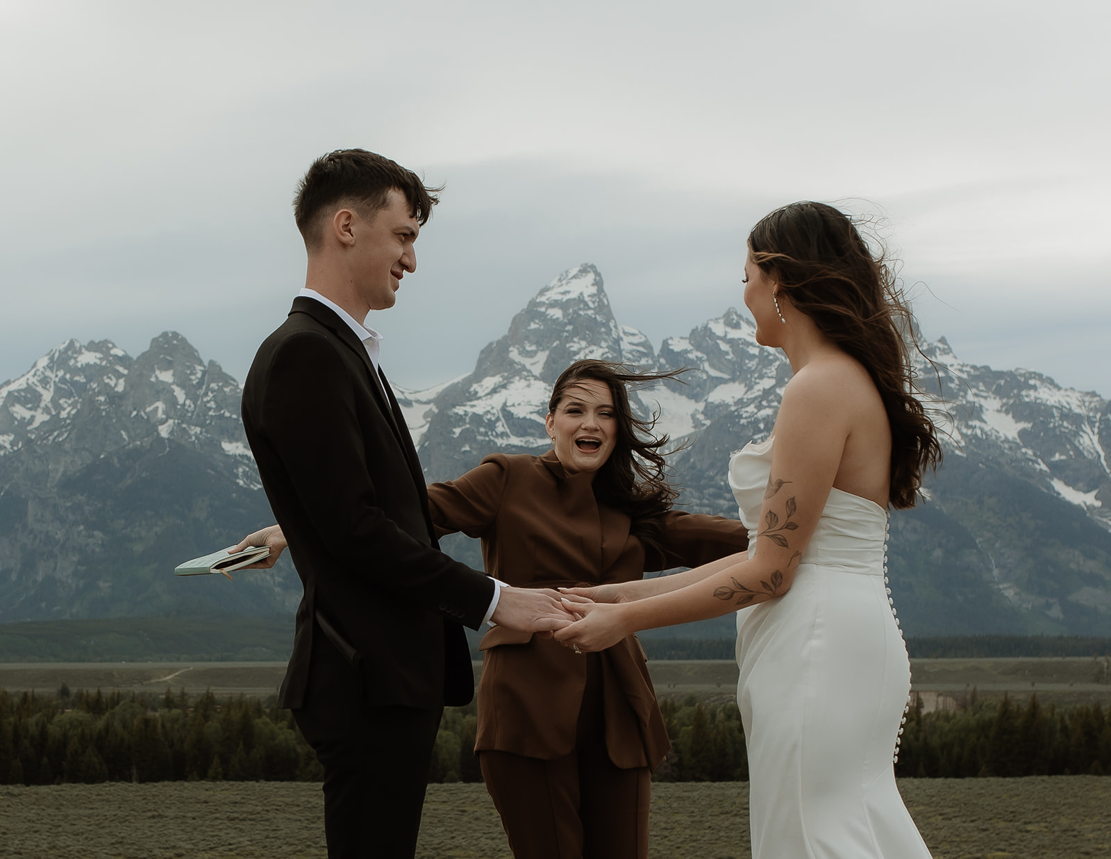 A couple stands facing each other holding hands during a wedding ceremony outdoors, with a person in the middle reading from a book. Snow-capped mountains are visible in the background for their elopment at Glacier View Turnout