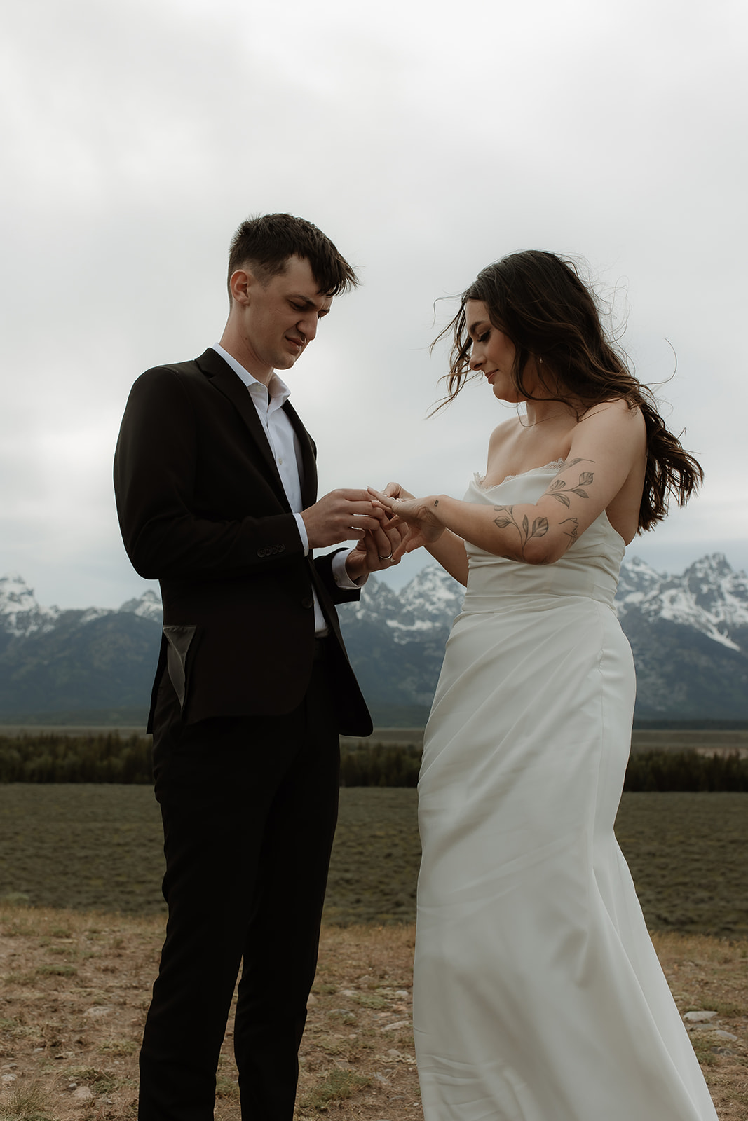 A couple stands facing each other holding hands during a wedding ceremony outdoors, with a person in the middle reading from a book. Snow-capped mountains are visible in the background for their elopment at Glacier View Turnout