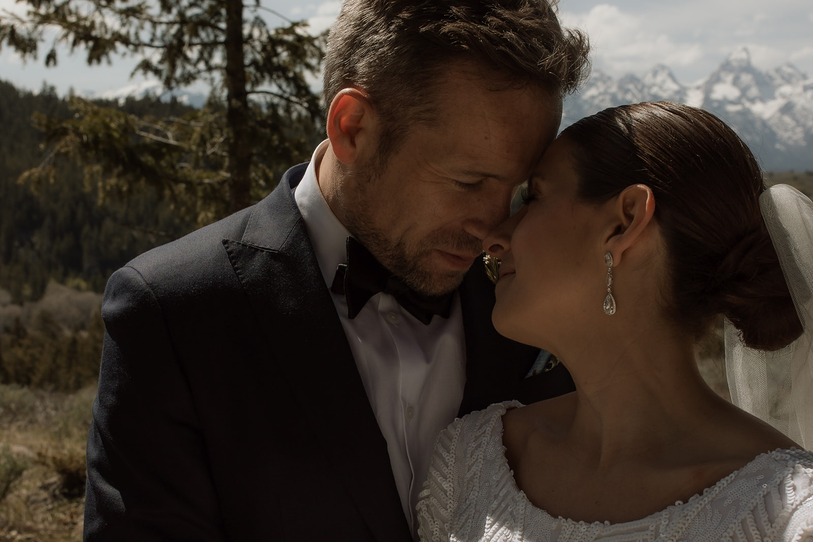 A bride and groom gaze at each other while standing outdoors with snow-capped mountains in the background. The bride holds a bouquet of flowers 