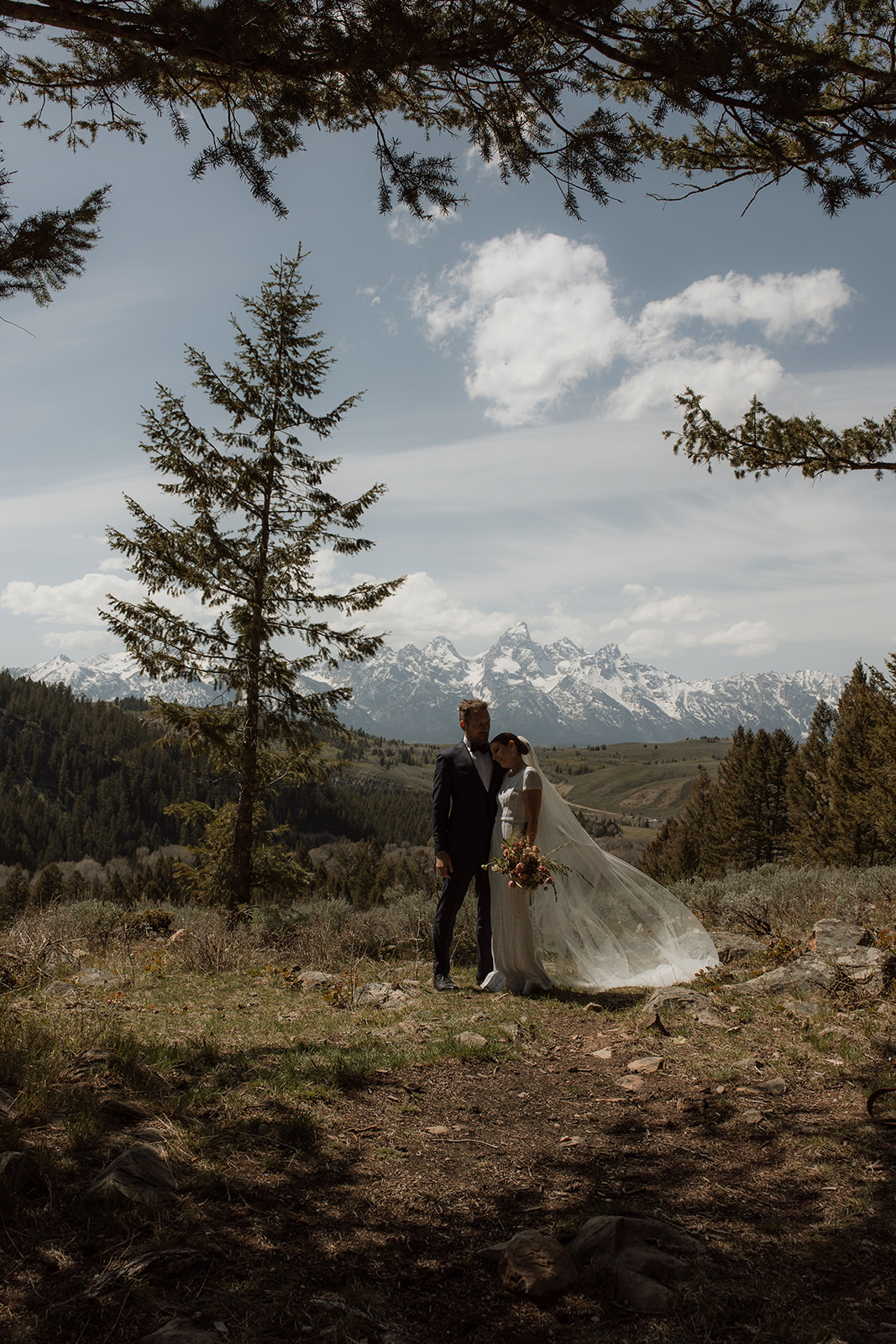 A bride and groom gaze at each other while standing outdoors with snow-capped mountains in the background. The bride holds a bouquet of flowers