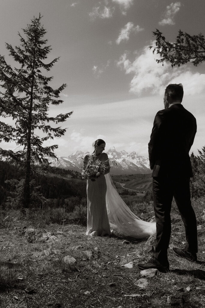 A bride and groom gaze at each other while standing outdoors with snow-capped mountains in the background. The bride holds a bouquet of flowers 