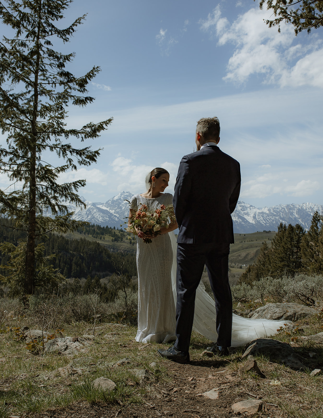 A bride and groom gaze at each other while standing outdoors with snow-capped mountains in the background. The bride holds a bouquet of flowers 