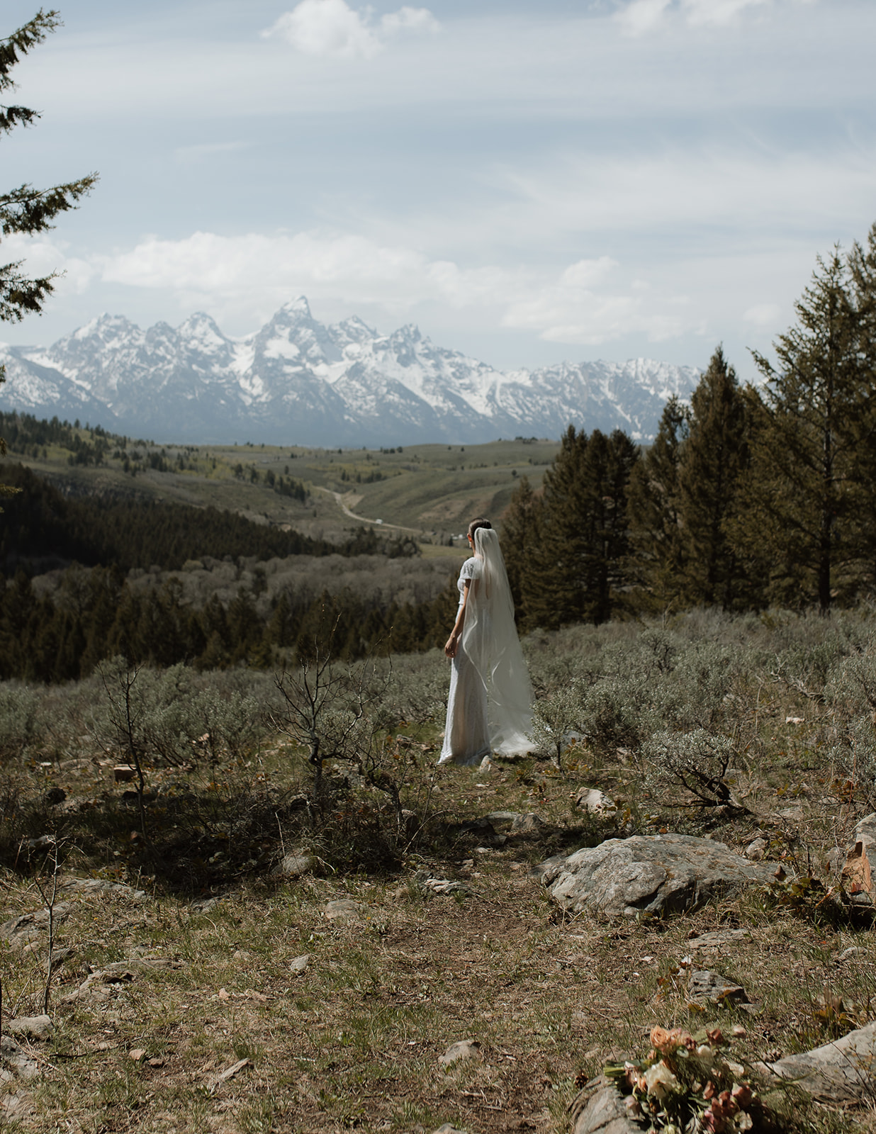 A bride and groom gaze at each other while standing outdoors with snow-capped mountains in the background. The bride holds a bouquet of flowers 