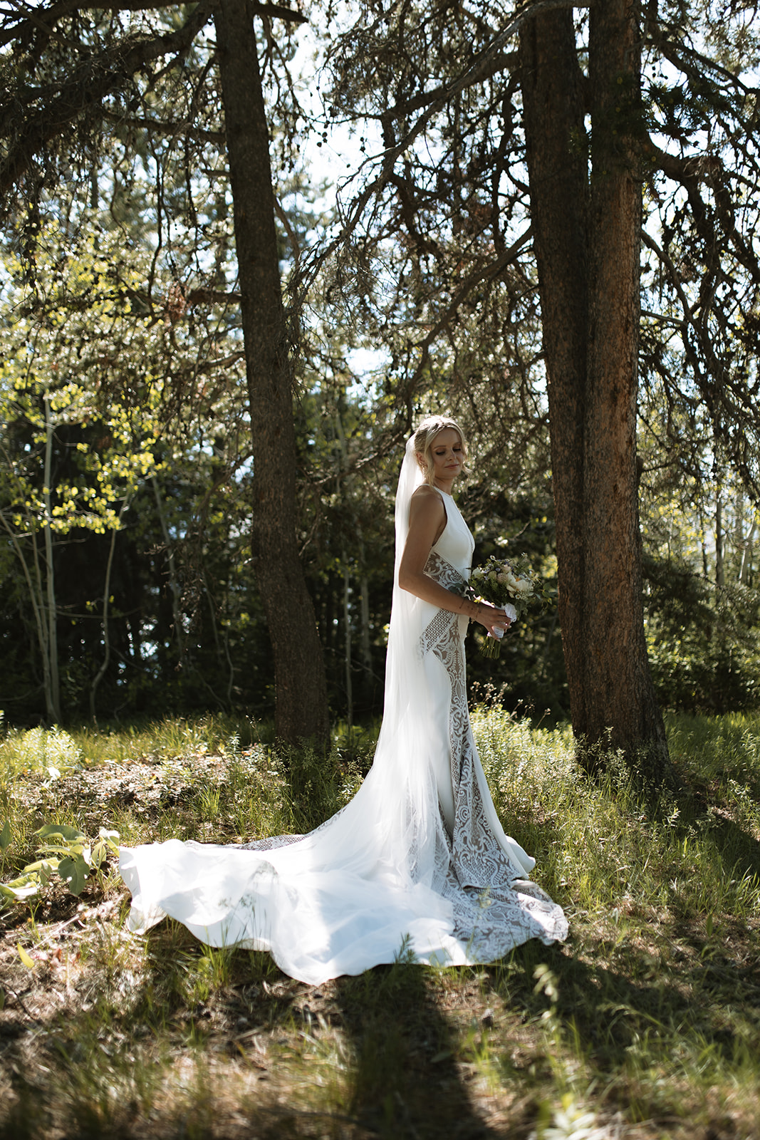 A bride and groom stand outdoors by a tree. The groom, in a black suit, reads from a piece of paper while the bride, in a white dress with a long veil, stands facing him at their JACKSON HOLE ELOPEMENT at Colter Bay