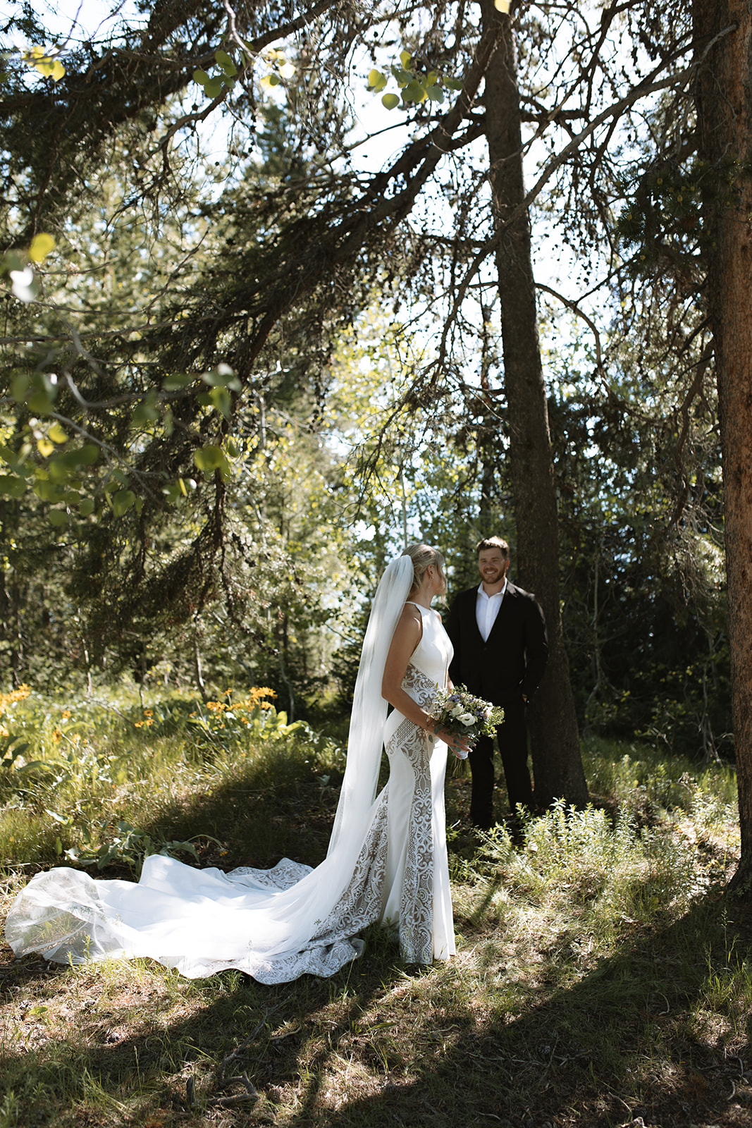 A bride and groom stand outdoors by a tree. The groom, in a black suit, reads from a piece of paper while the bride, in a white dress with a long veil, stands facing him at their JACKSON HOLE ELOPEMENT at Colter Bay