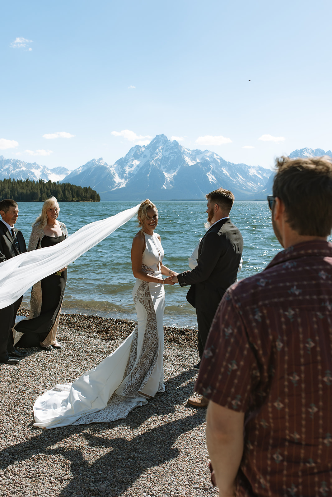 A couple exchanges rings in a wedding ceremony by a body of water, officiated by a woman holding a book. The bride is in a white dress with a veil, and the groom is in a dark suit at an elopement at Colter Bay in Jackson Hole 