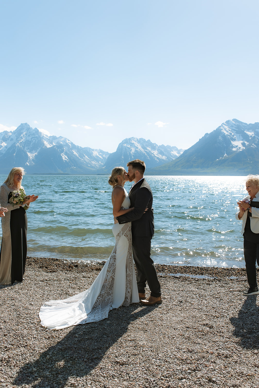 A couple exchanges rings in a wedding ceremony by a body of water, officiated by a woman holding a book. The bride is in a white dress with a veil, and the groom is in a dark suit at an elopement at Colter Bay in Jackson Hole 