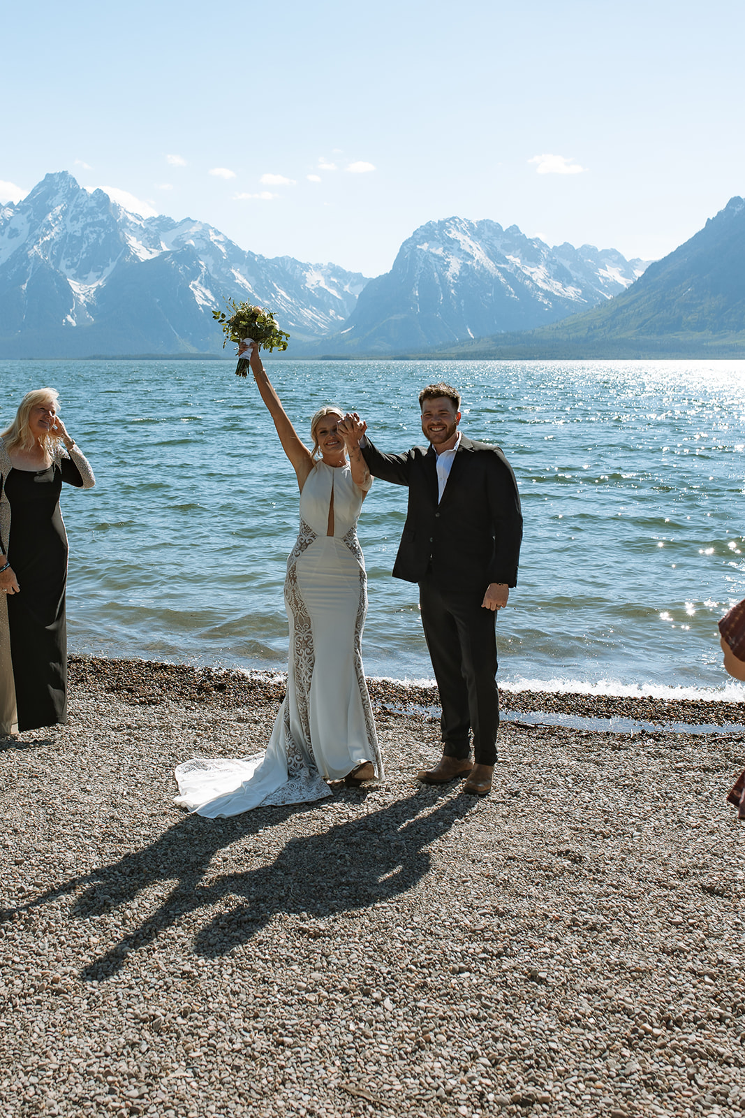 A couple exchanges rings in a wedding ceremony by a body of water, officiated by a woman holding a book. The bride is in a white dress with a veil, and the groom is in a dark suit at an elopement at Colter Bay in Jackson Hole