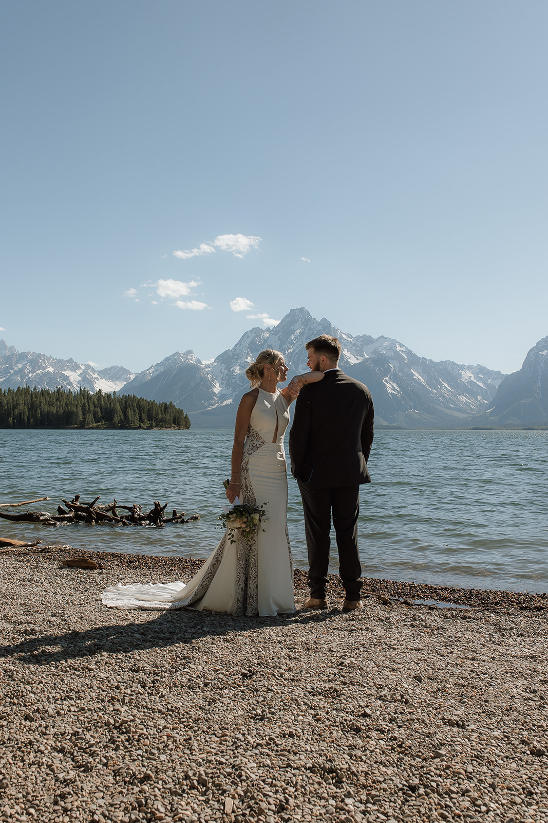 A couple stands by a lake, facing away, with mountains and trees in the background under a partly cloudy sky. She is wearing a white dress, and he is in a black suit for their elopement at jackson hole