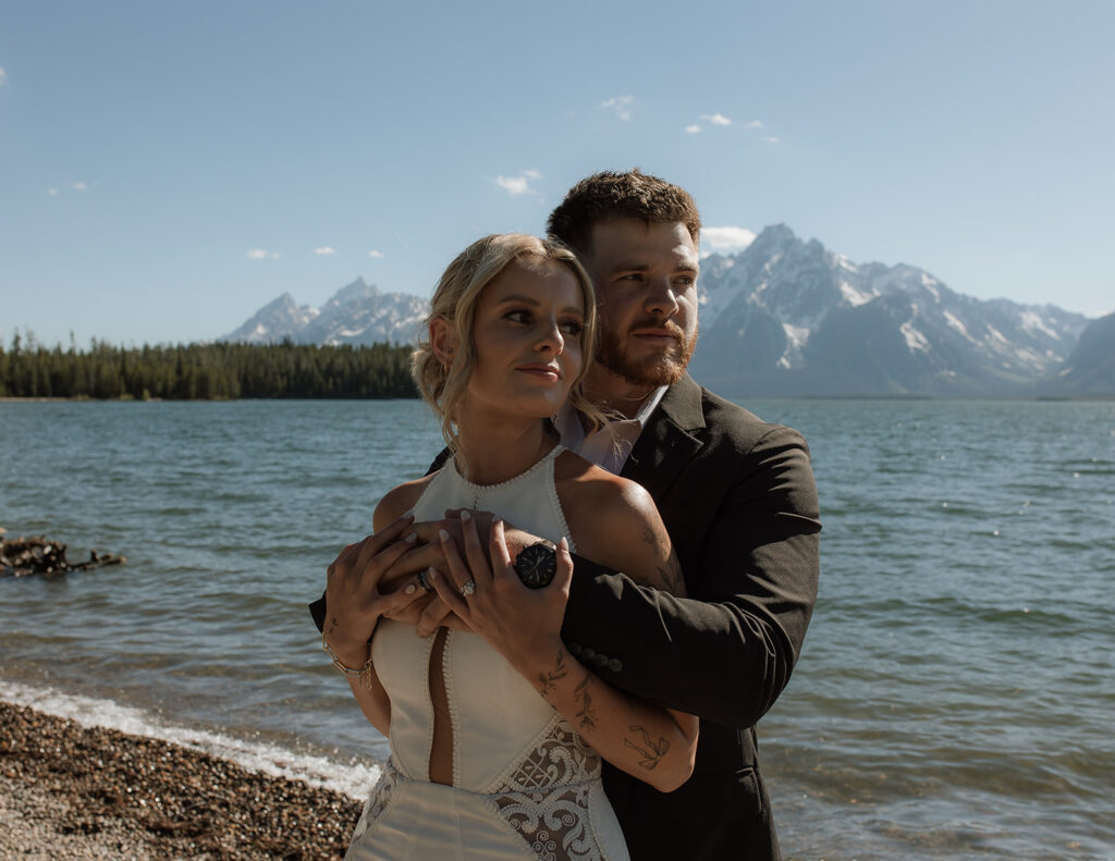 A couple stands on a rocky beach with a mountainous lake backdrop. The man embraces the woman from behind as they both look into the distance.